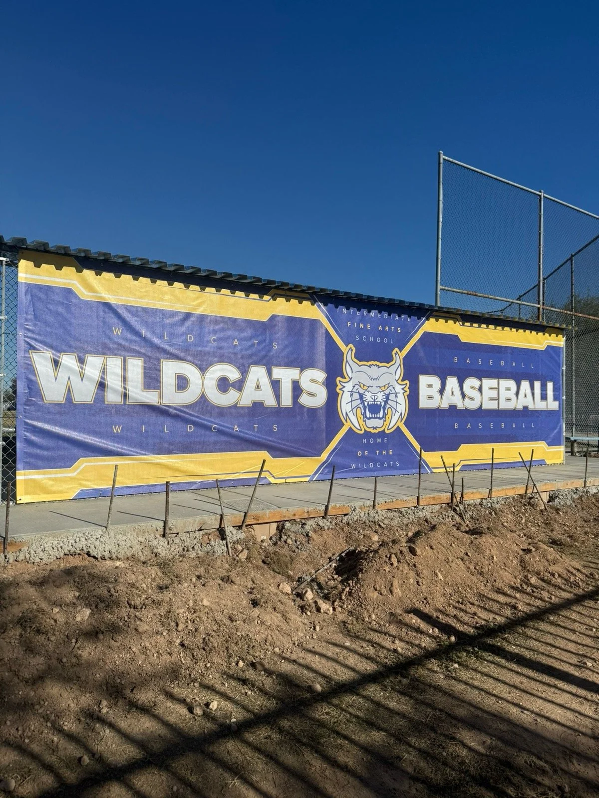 A large blue and yellow banner on a chain-link fence promotes the Wildcats baseball team at a school. The banner features a wildcat's face and phrases like "Wildcats" and "Home of the Wildcats," with a clear sky above and a dirt area in front.