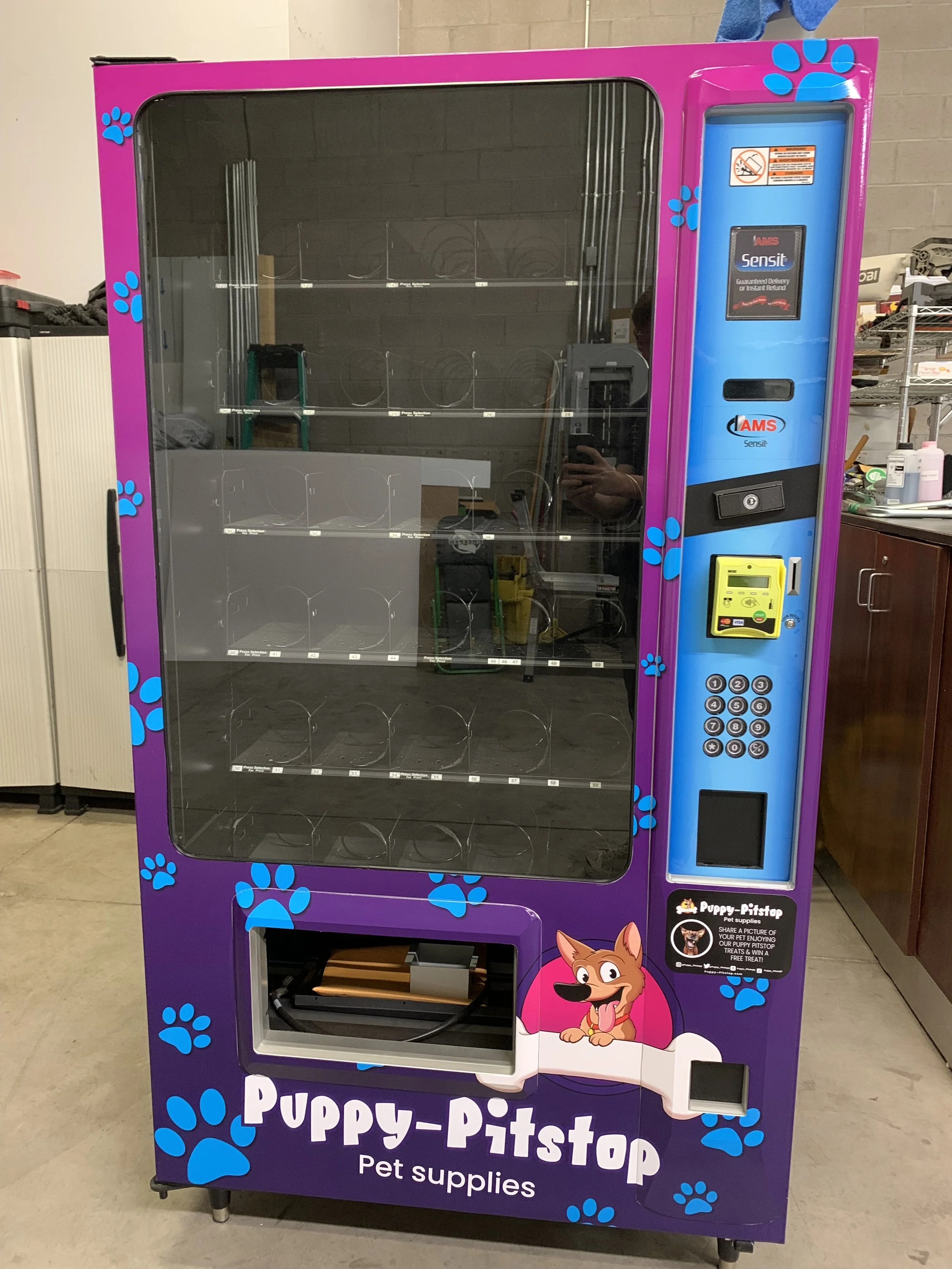 A vending machine designed for pet supplies with a purple and blue wolf theme, featuring paw print decorations. The machine has a clear front showing empty shelves, a card reader, a keypad, and a small display screen.