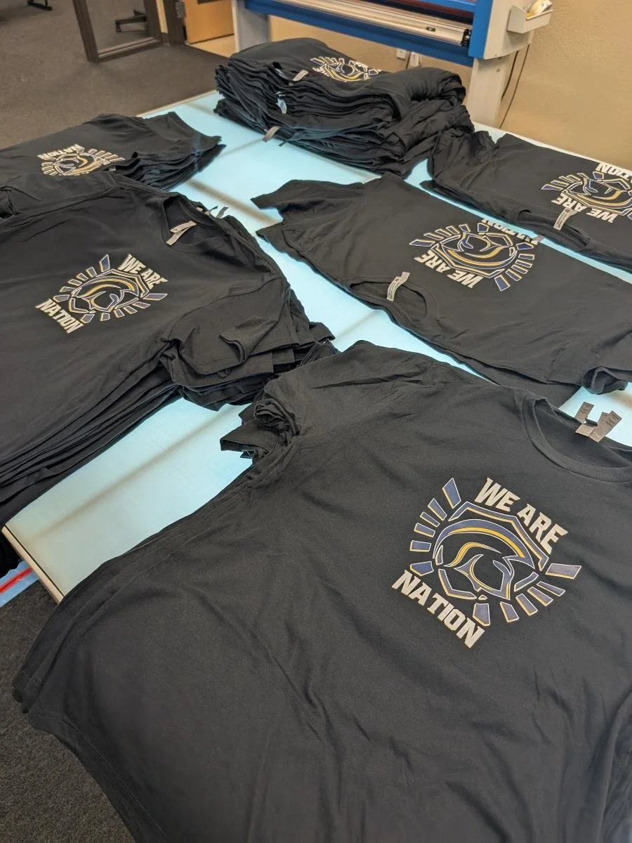 Multiple black T-shirts with a logo that says "We Are Nation" and features a stylized eagle or hawk, laid out on a table. The T-shirts appear to be prepared for distribution or sale.