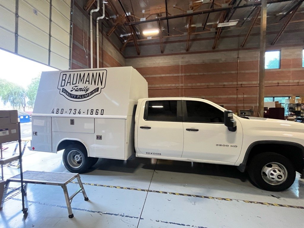 White service utility truck with 'Baumann Family Quality Plumbing' logo and contact number parked inside a warehouse.
