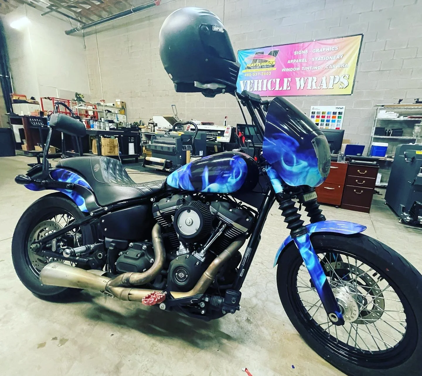 Motorcycle with custom blue flame paint job, parked indoors in a workshop or garage, with various tools and equipment in the background, including a large helmet placed on the front of the motorcycle.