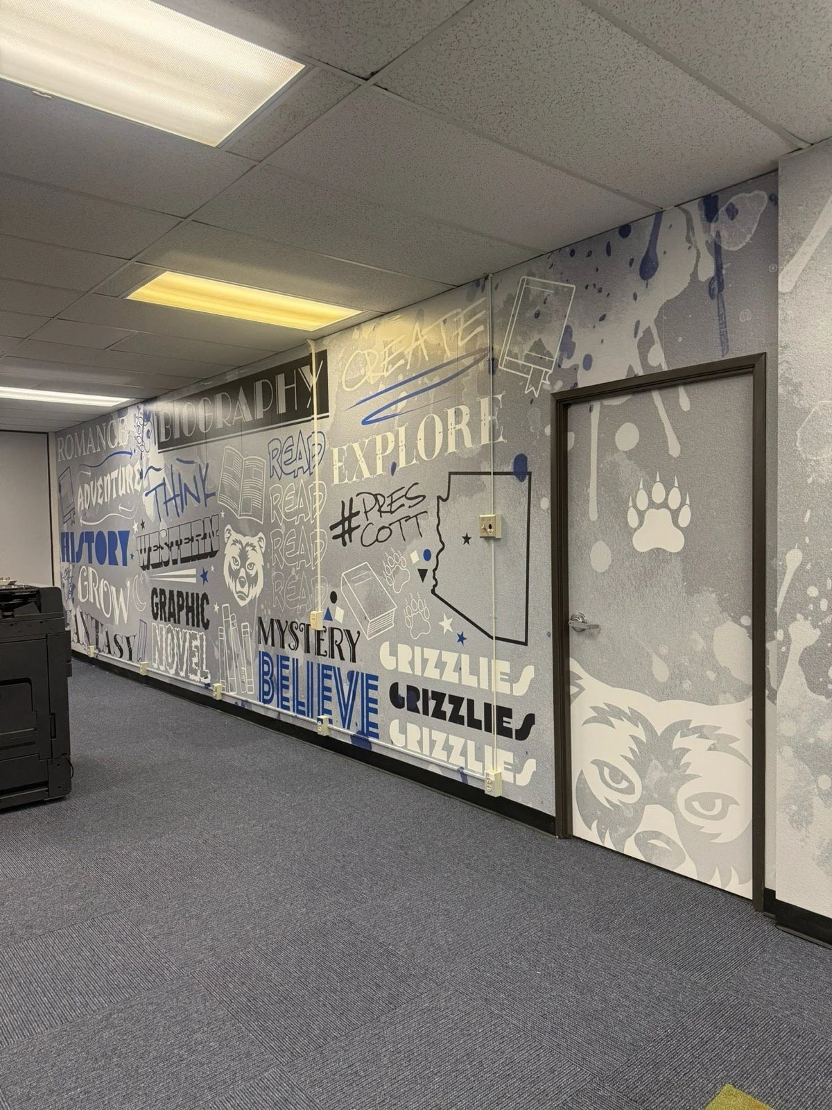 Decorative wall in an office with words like 'biography,' 'explore,' 'read,' 'believe,' and illustrations including a bear face, paw prints, and text such as '#prescott' and 'grizzlies' in blue, black, and white.