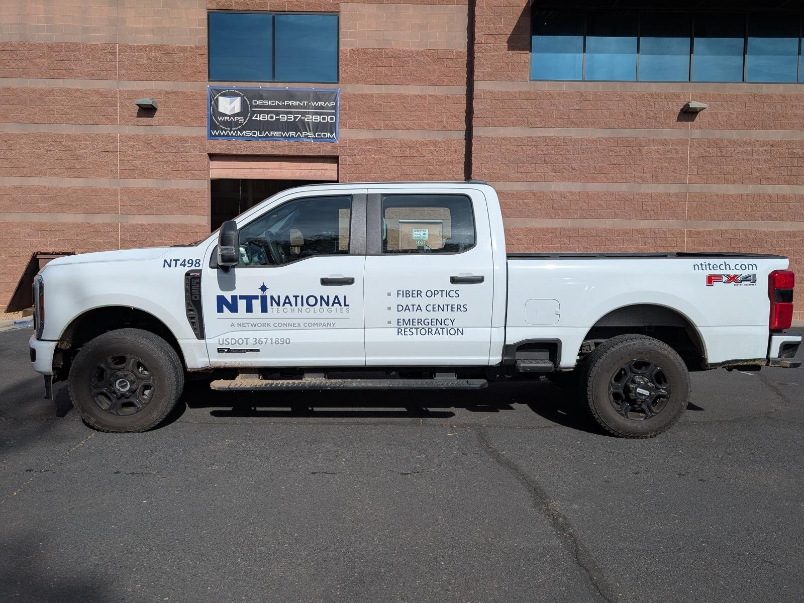 White pickup truck with logos and text for NTI National Technologies, parked in front of a brick building with a blue sign.