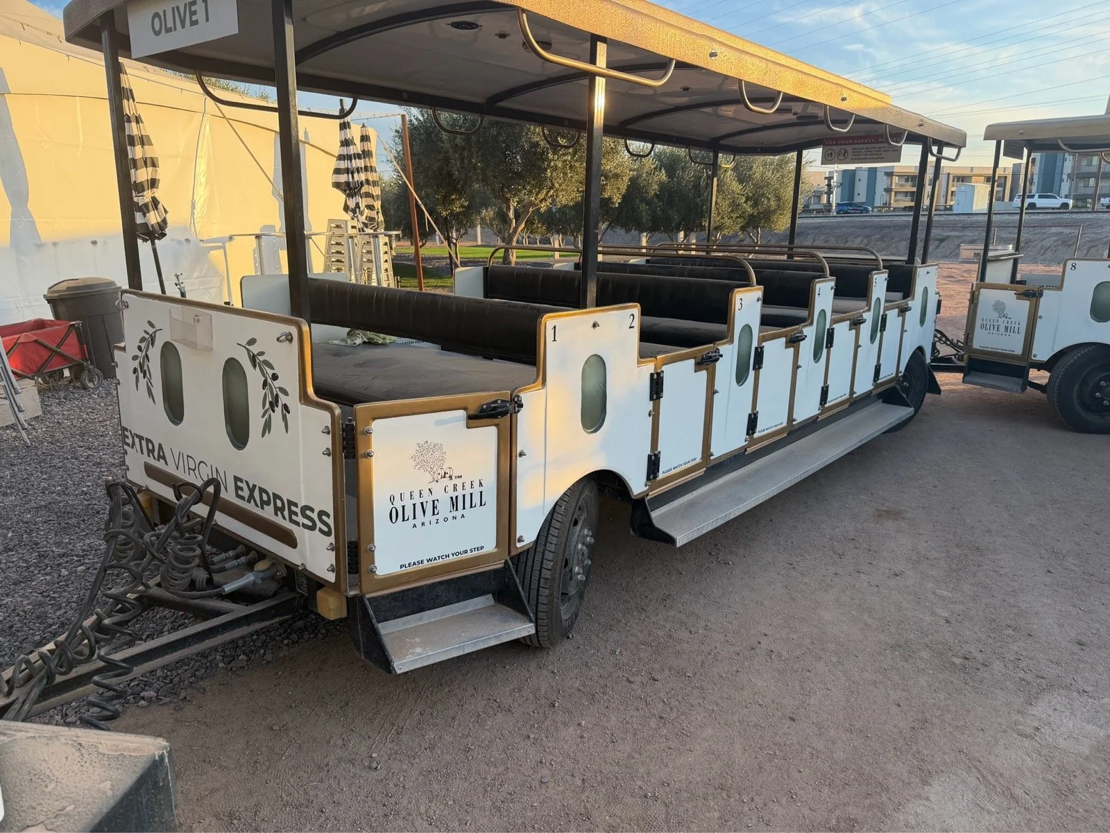 A white tram decorated for a wine tour, with black cushioned seats and striped umbrellas, marked as Extra Virgin Express at Queen Creek Olive Mill, Arizona.
