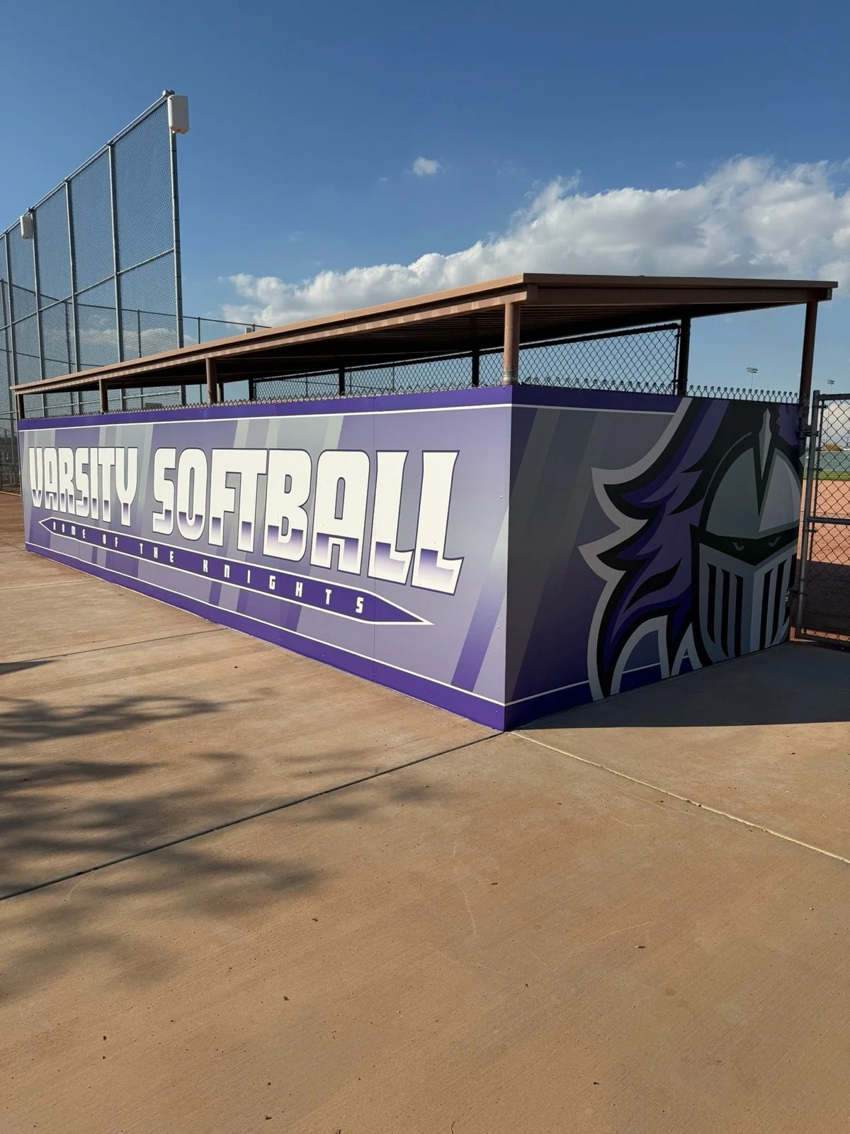 A purple and gray sign on a fence reads "Varsity Softball - Home of the Knights" with a knight's helmet logo.