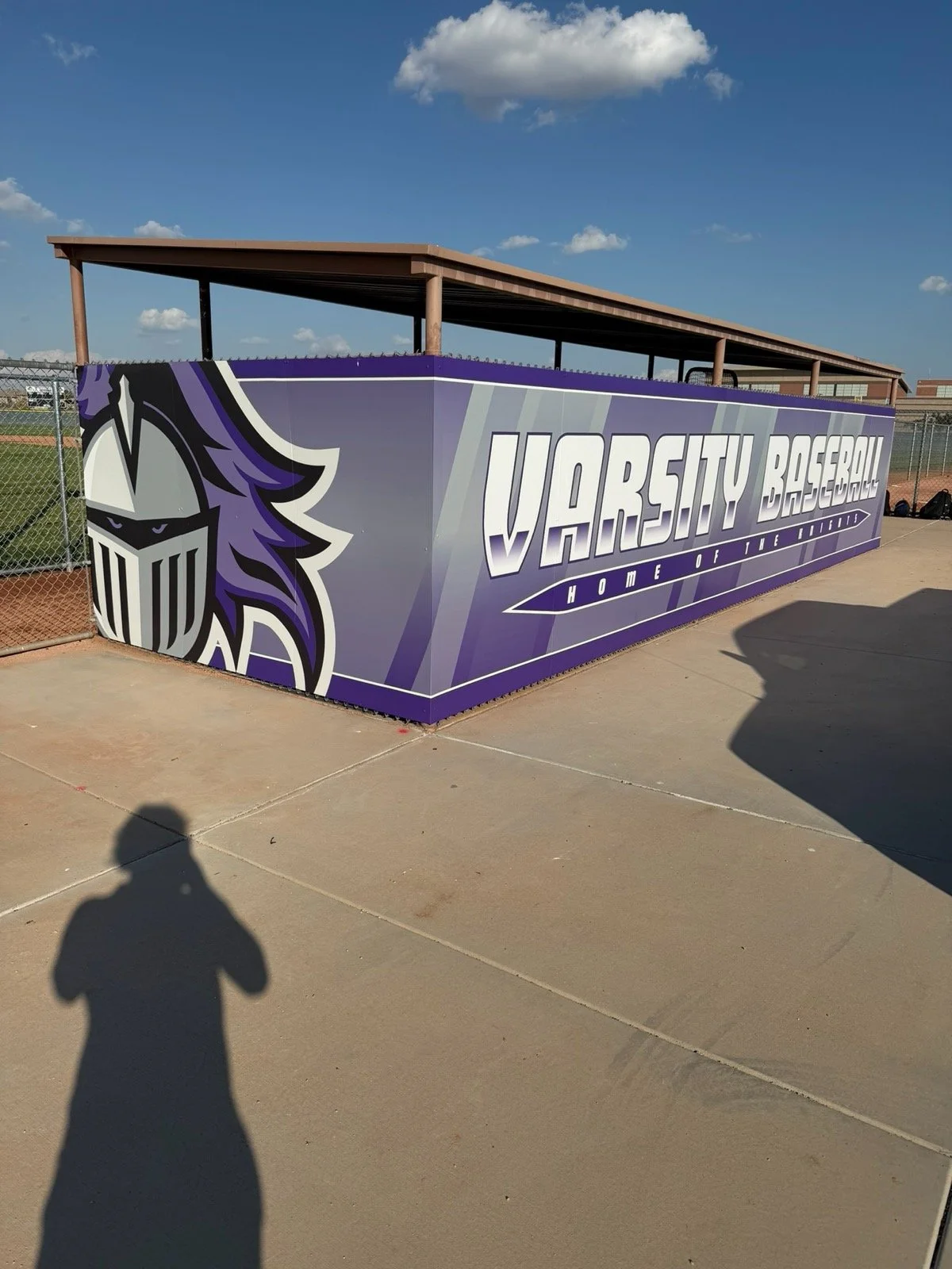 A purple and white sign with the text 'VARSITY BASEBALL HOME OF THE TIGERS' and a tiger mascot logo, attached to a chain-link fence at a baseball field under a blue sky with clouds.