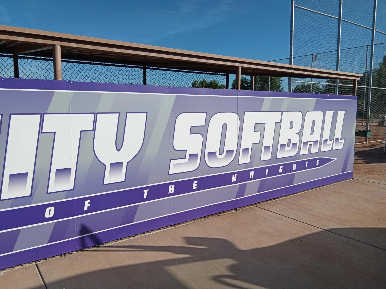 A sign on a sports field that reads 'LILY SOFTBALL' with the subtitle 'OF THE KNIGHTS,' with a blue sky and chain-link fence in the background.