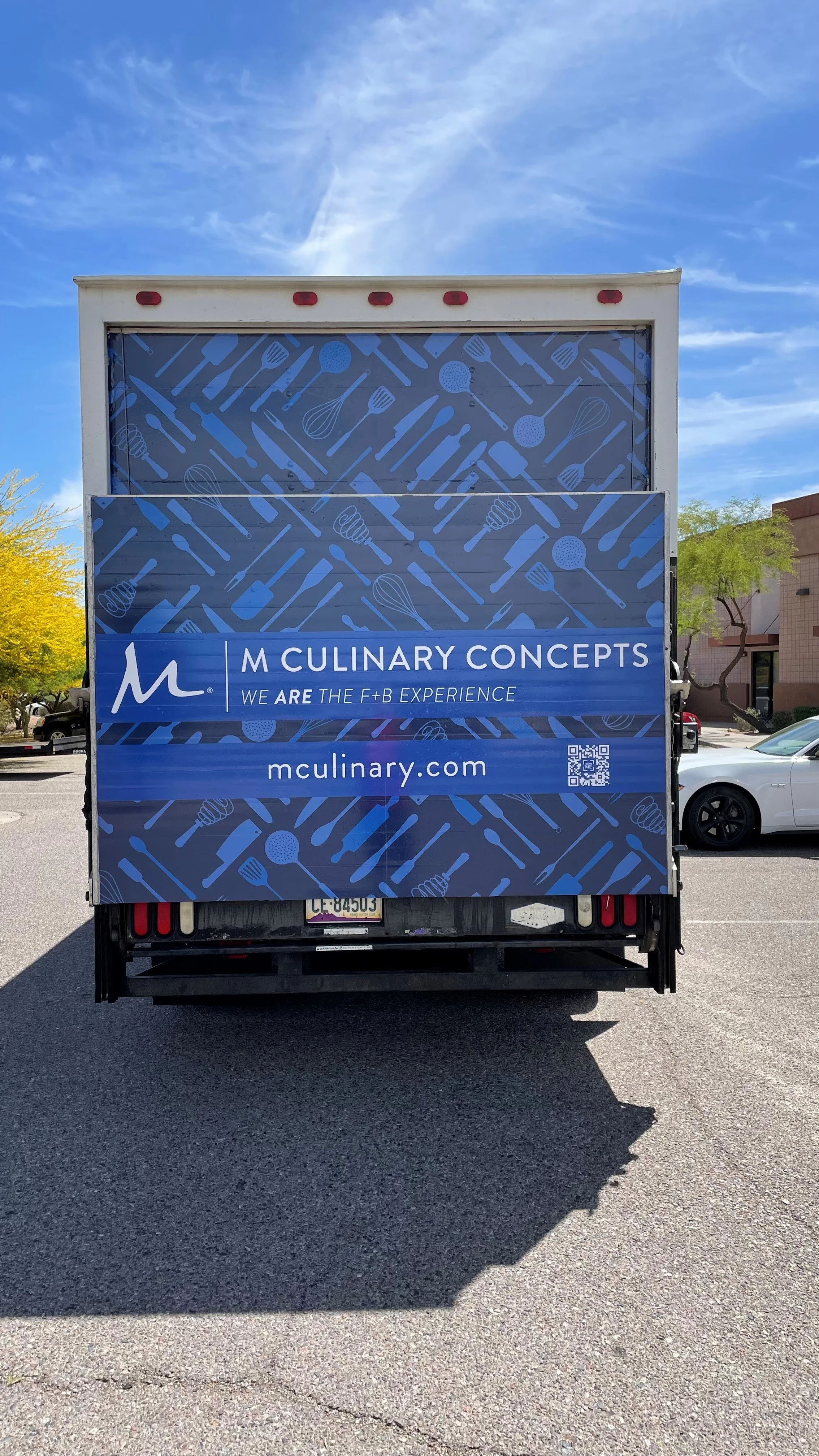 Back of a food truck with blue advertising for M Culinary Concepts, featuring kitchen utensils and a QR code, parked in a lot with trees and a building in the background.