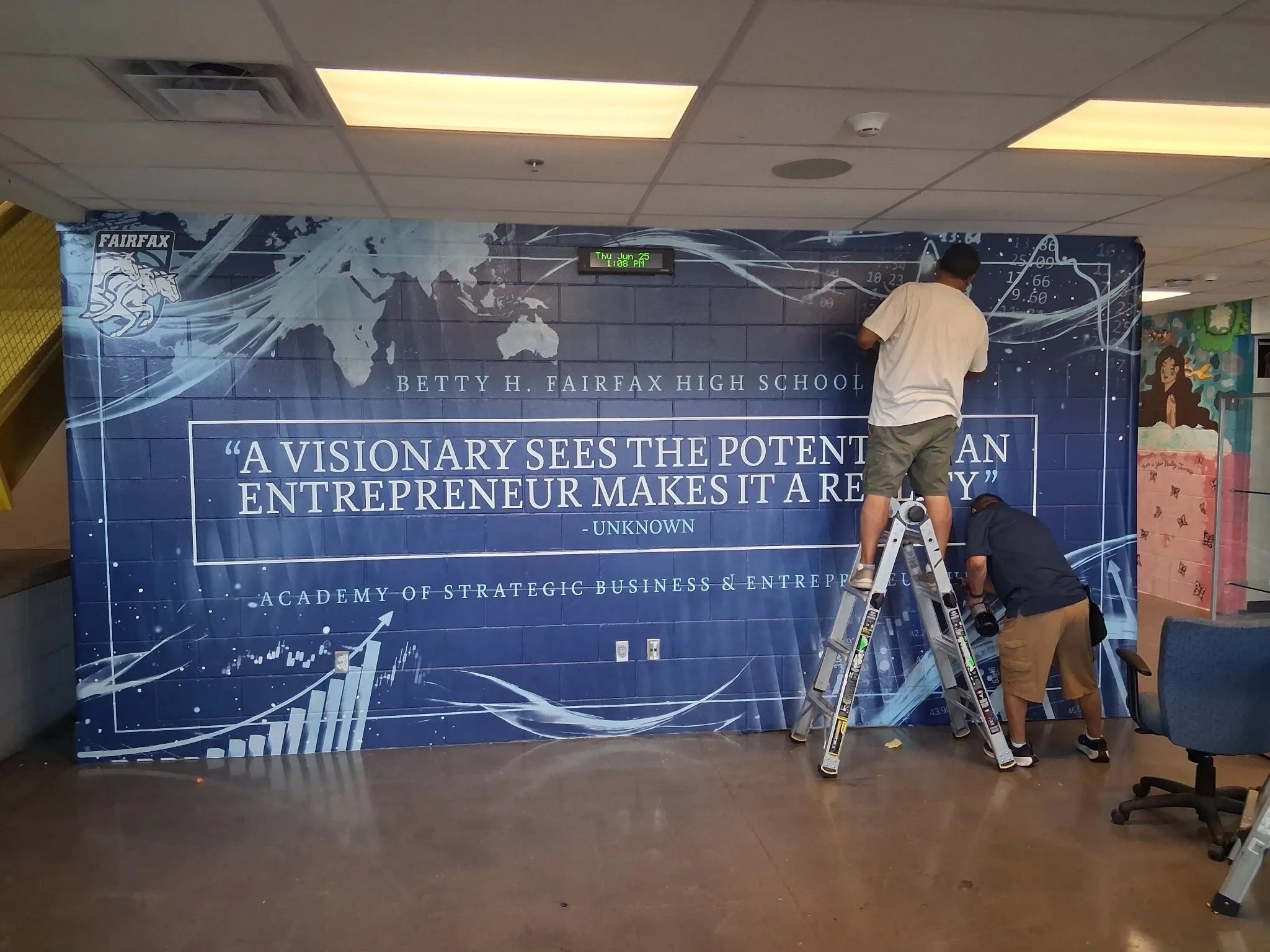 People in a hallway installing a large blue banner for Betty H. Fairfax High School that features a quote about visionary leadership and a world map. One person on a ladder is attaching the banner, while another crouches for assistance.