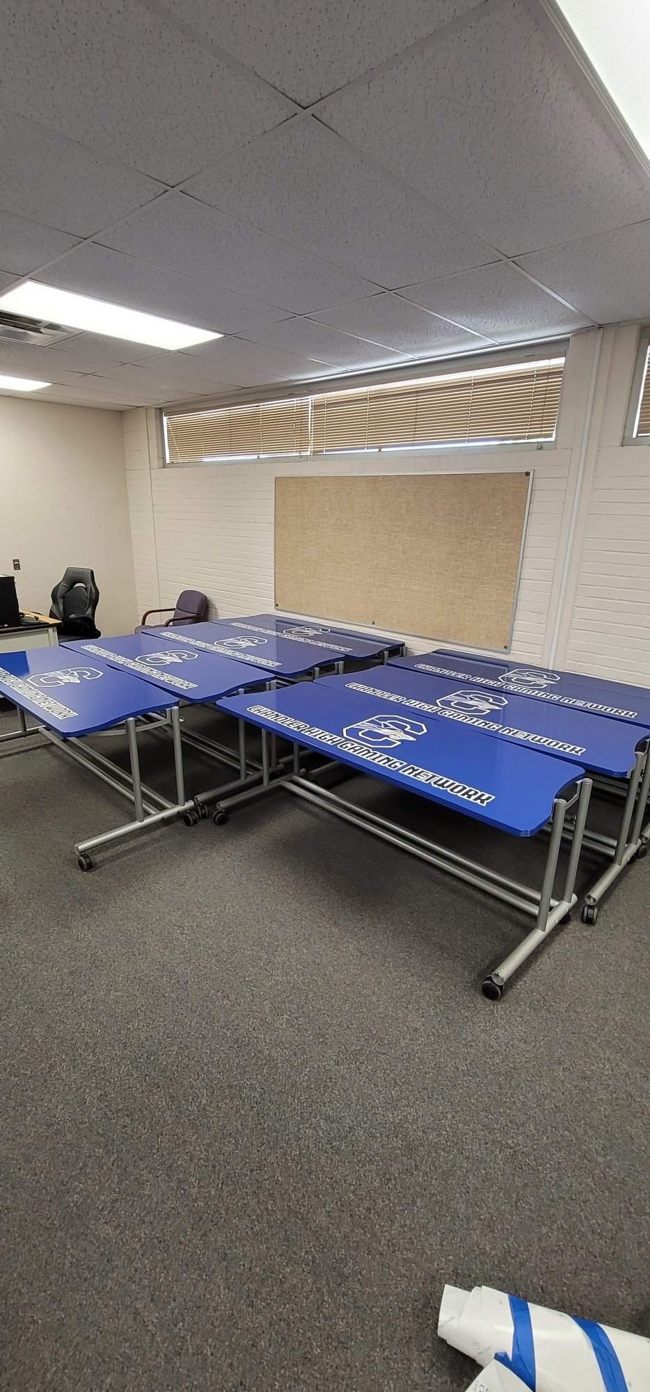 A room set up for gaming events with blue tables featuring the 'Ganderhatch Gaming Network' logo, a whiteboard on the wall, office desk and chair, and window blinds.