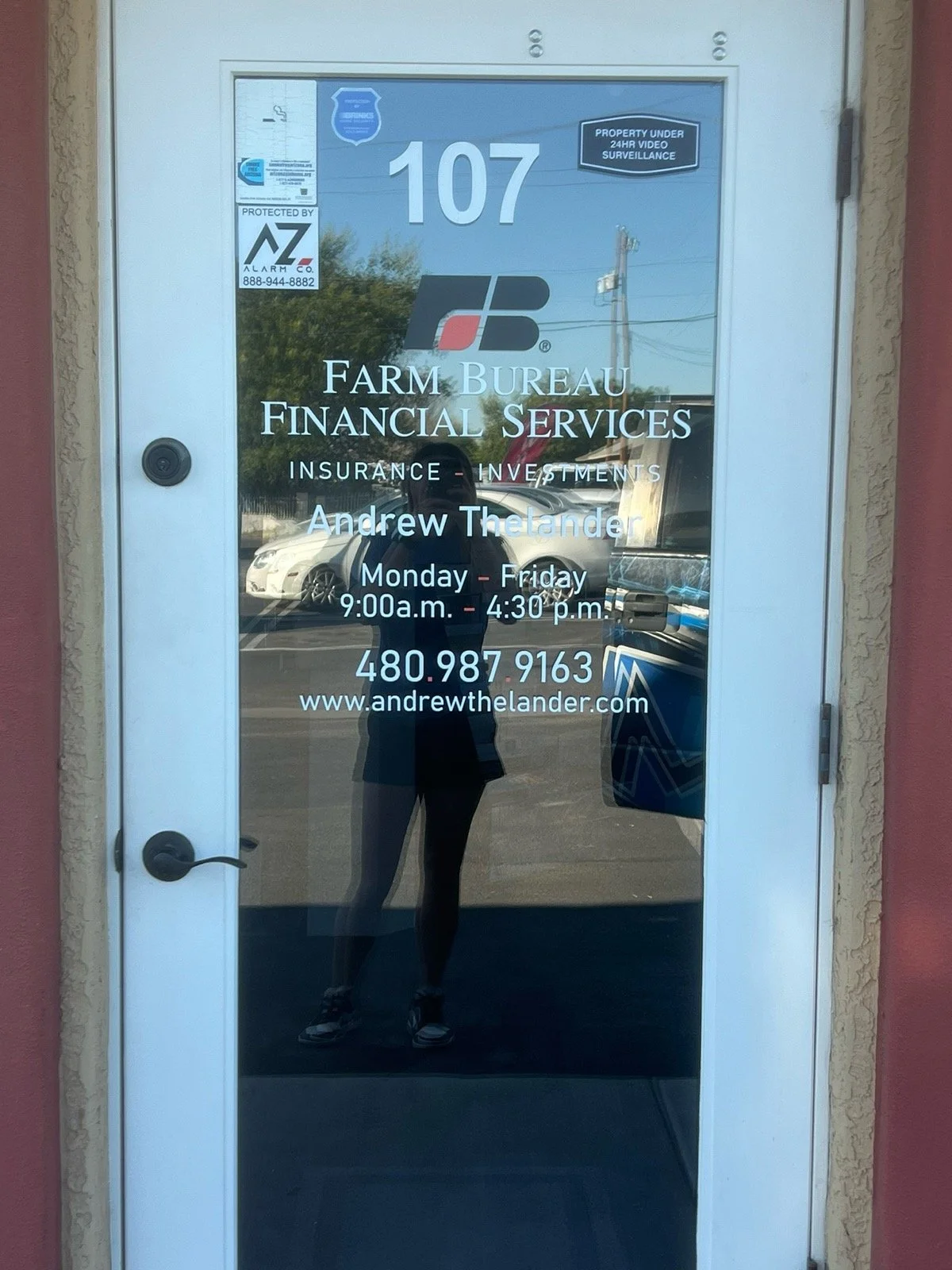 Glass door with business sign for Farm Bureau Financial Services, showcasing contact information, operating hours, and a reflective background with parked cars and a person taking the photograph.