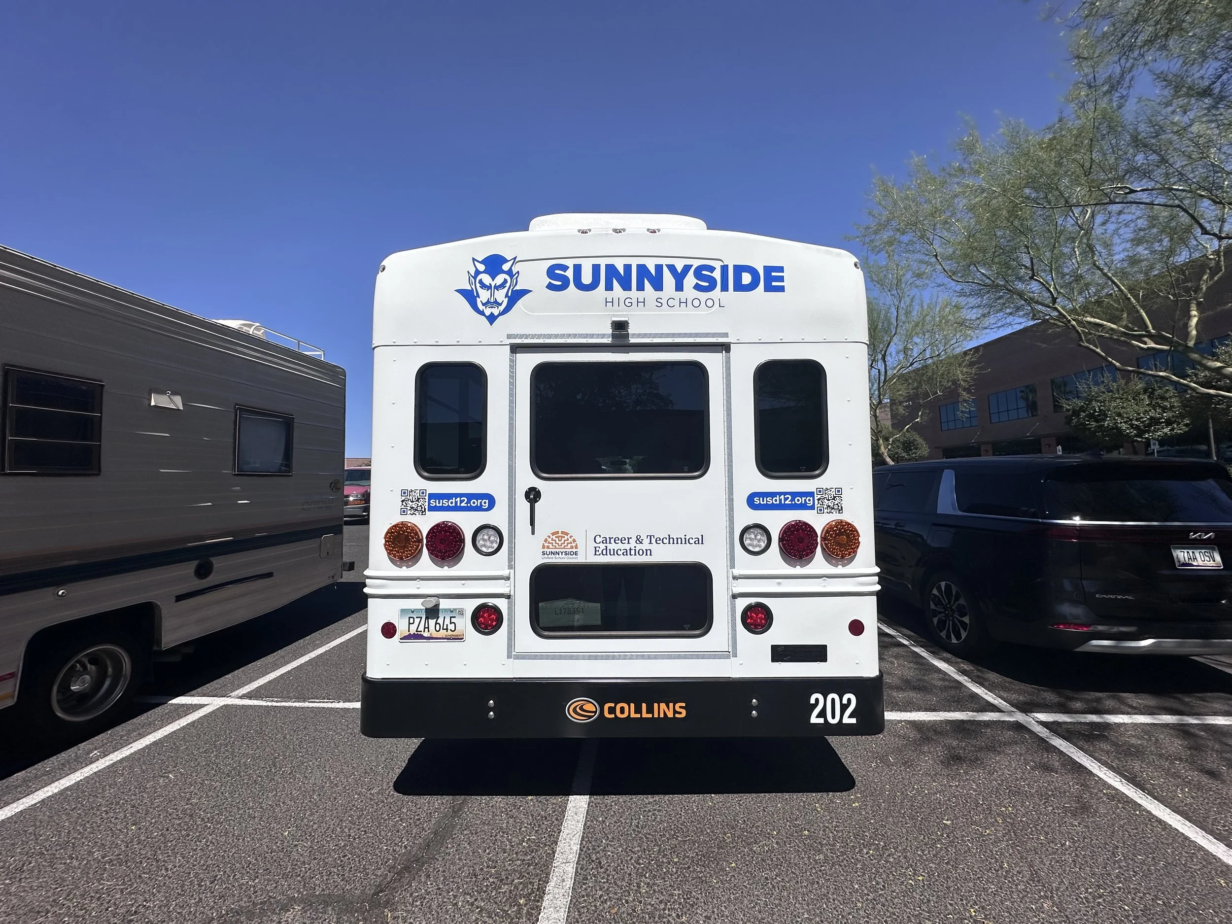 The back of a white bus with the logo and name of Sunnyside High School, including their mascot and website, parked in a lot with other vehicles and buildings visible in the background under a clear blue sky.