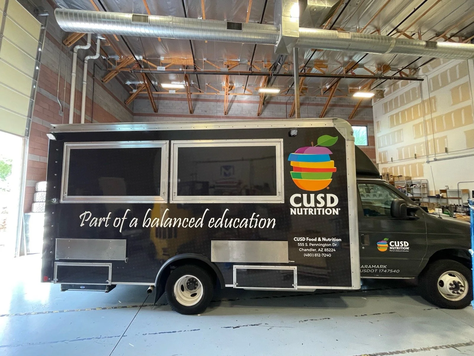Black food truck with colorful logo and text, parked inside a warehouse. Logo depicts a colorful apple with layers, and includes the text 'CUSD NUTRITION.' The side of the truck has the slogan 'Part of a balanced education.' Additional information in