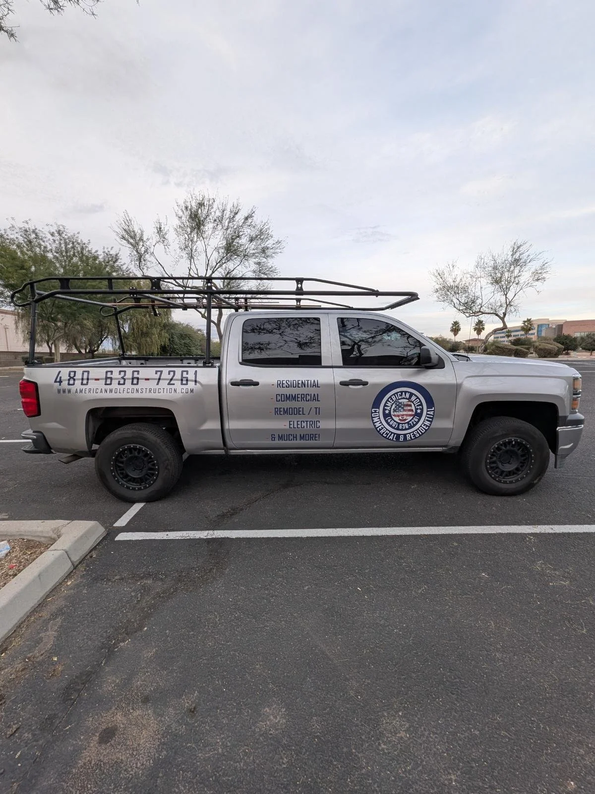 Silver pickup truck parked in a parking lot with company information and services printed on the side, including phone number and website, with trees and a cloudy sky in the background.