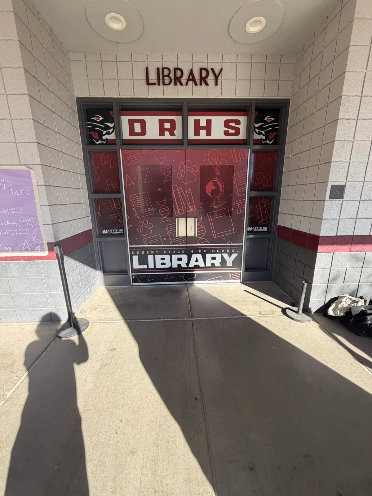 Entrance to Desert Ridge High School library with glass doors and red and white signage, including illustrations of books and lightbulb, and a purple board to the left with schedule information.