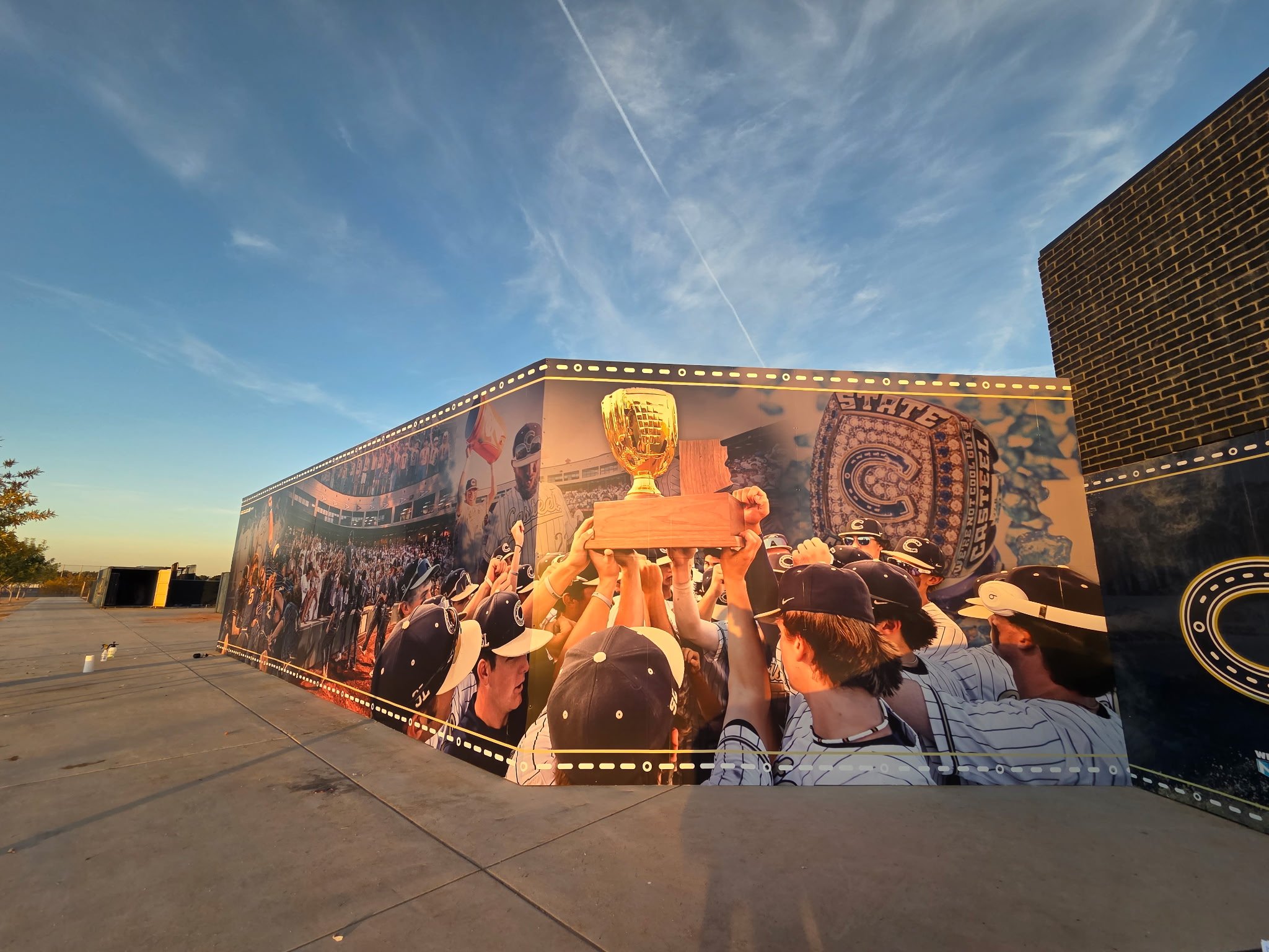 A large outdoor wall mural depicting a sports team celebrating with a trophy, surrounded by fans in team jerseys and hats, under a blue sky with some clouds and contrails.