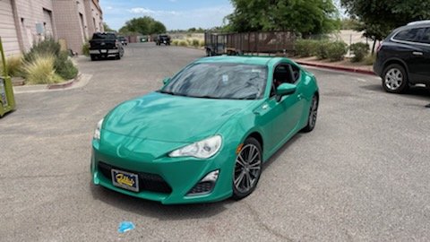 A teal-colored sports car is parked in a lot near a sidewalk, with other cars and buildings in the background.