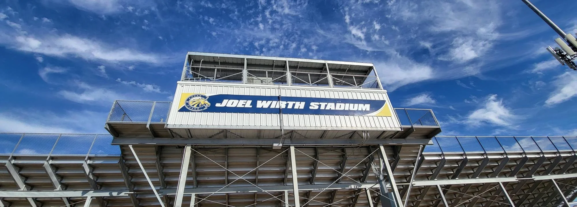 View of Joël Wirth Stadium's bleachers and press box against a blue sky with scattered clouds.