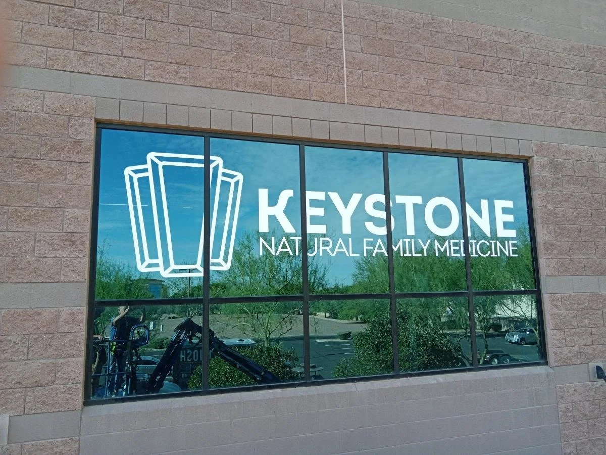 Storefront window with a logo of a stack of books and the text "KEYSTONE NATURAL FAMILY MEDICINE." Reflection of parking lot and trees outside.