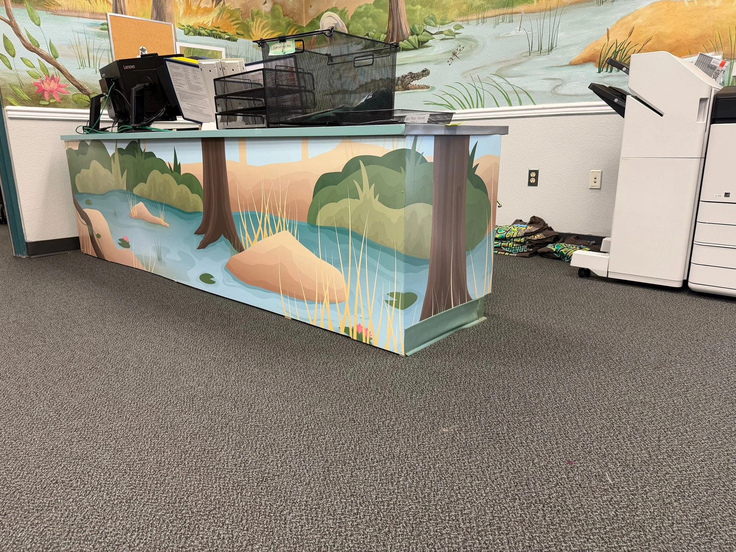 Reception desk with jungle-themed mural featuring a crocodile, trees, and plants, office supplies on the desk, and a white printer to the right.