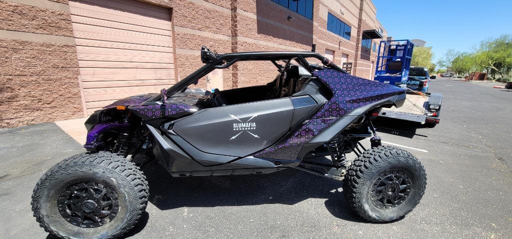 Off-road vehicle with large tires, black and purple body, parked in a lot near a brick building.