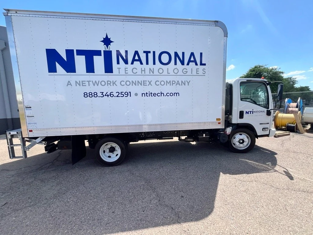A white truck with the logo and contact information for NTI Technologies, a network connectivity company, parked on a gravel lot with spool spools of cable on the background.