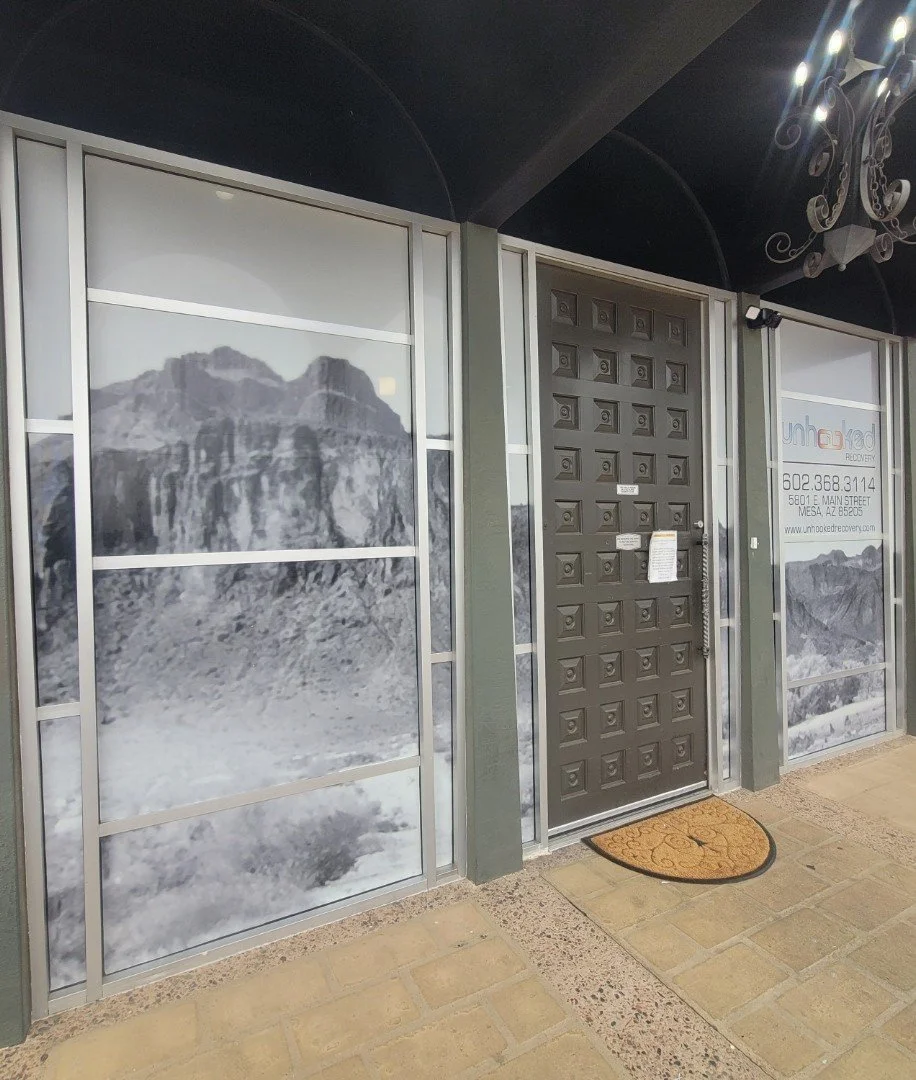 Storefront with mountain-themed decals, a solid black door with a pattern of small squares, and a window advertising Unhindered Recovery. There is a decorative chandelier overhead and a welcome mat in front of the door.