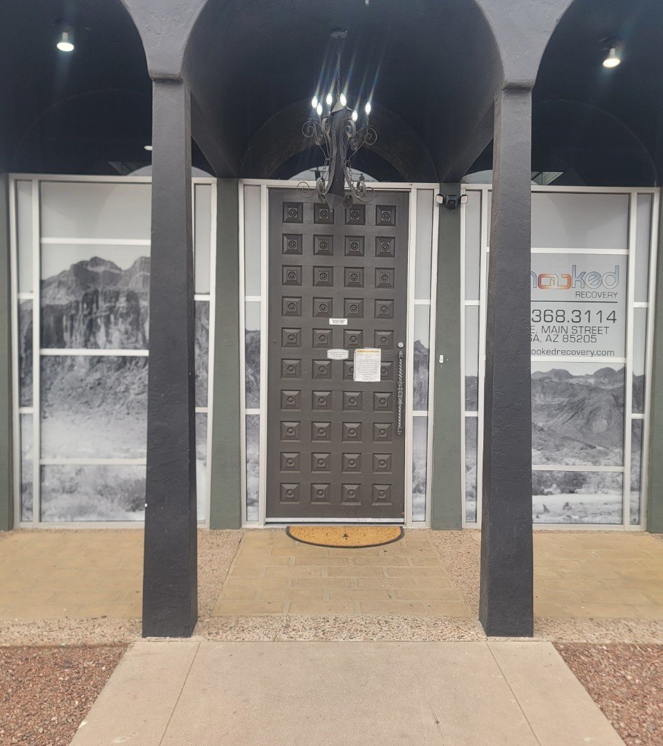 Black door with square patterns, surrounded by black and gray framing, with a semicircular orange doormat in front, leading to a recovery center called 'naked recovery' with a mountain landscape decal on the wall, located at 368 S. Main Street, Mesa,