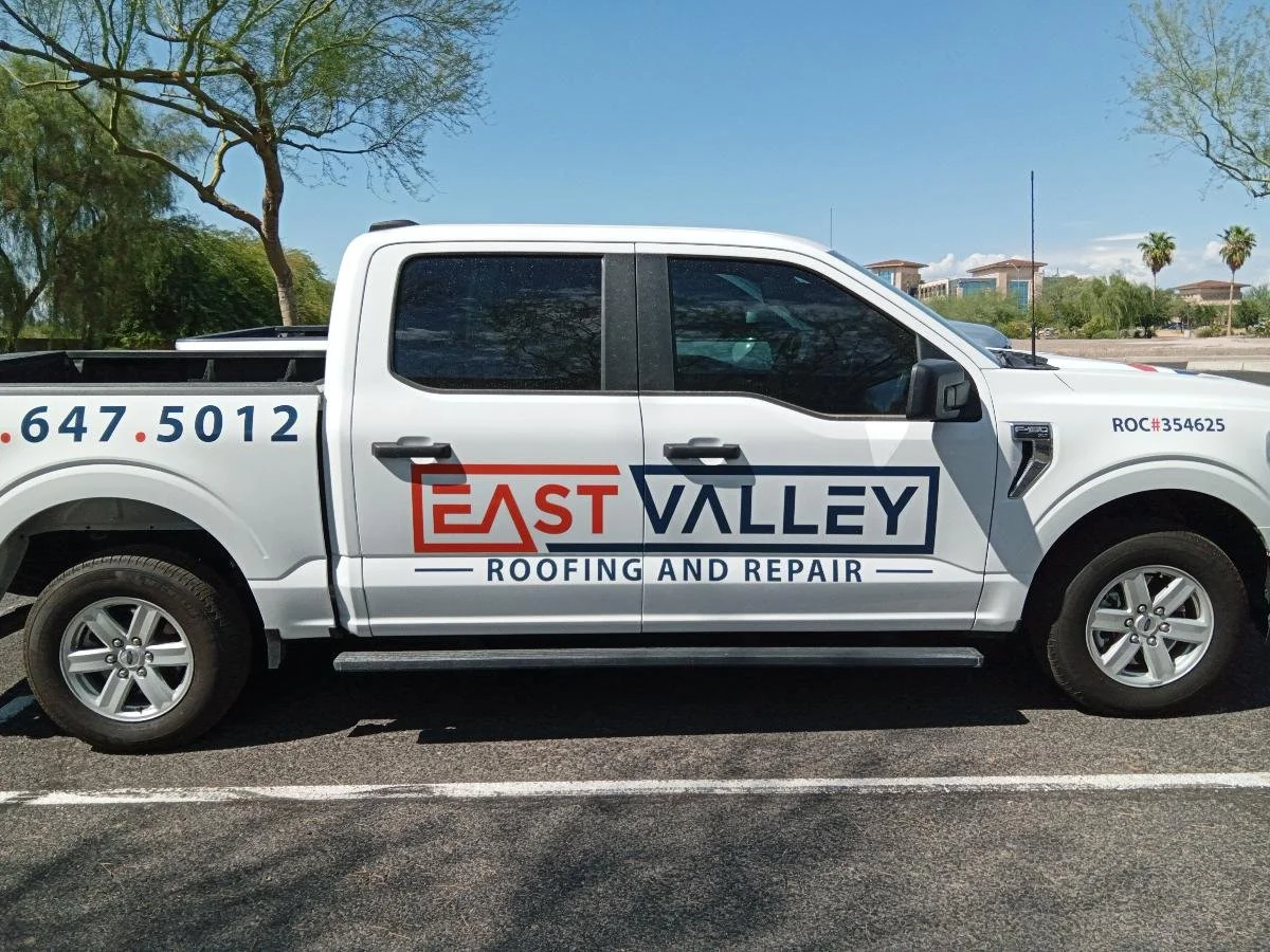 A white pickup truck with the logo and name 'East Valley Roofing and Repair' on the side. The truck is parked on a street with trees and buildings in the background.
