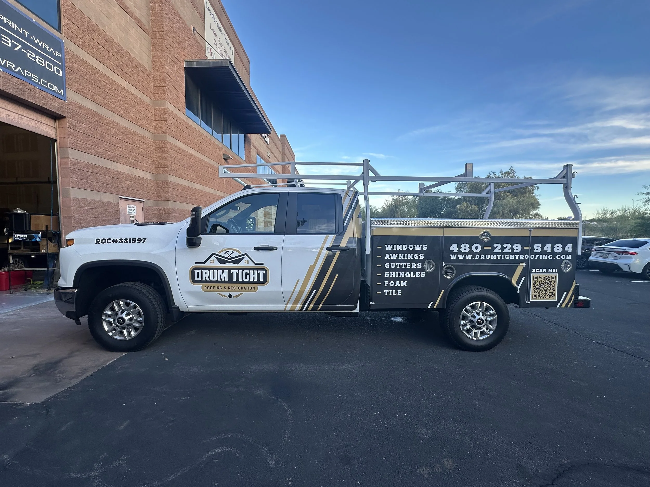 A white service truck with black and gold branding for Drum Tight Roofing & Restoration parked outside a building with a brick facade. The truck displays services such as windows, awnings, gutters, shingles, foam, and tile, along with contact informa