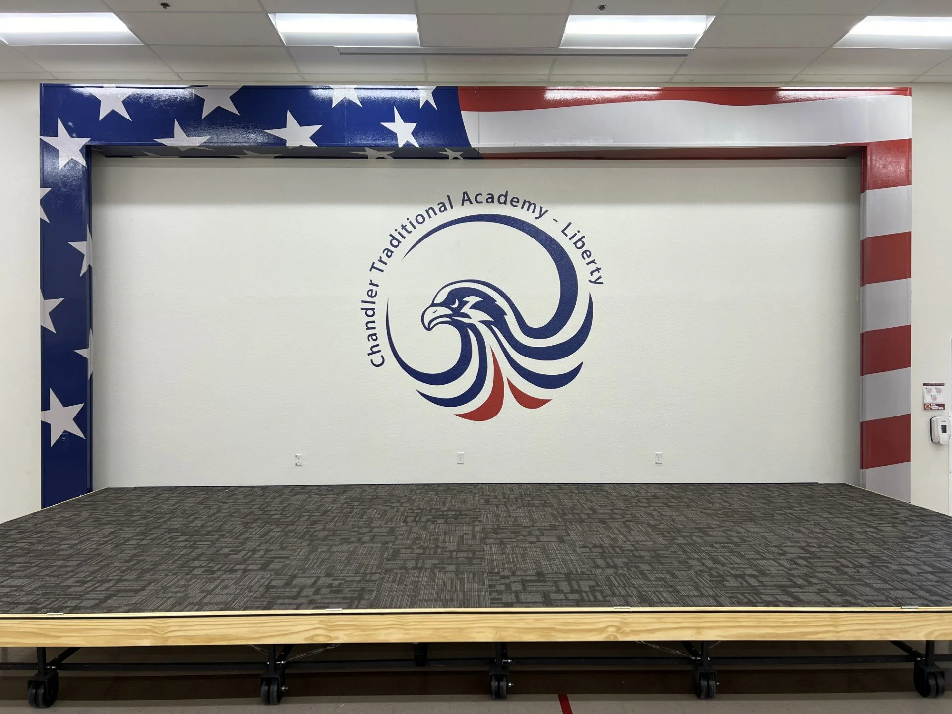 Interior of a room with a white wall and flag-themed decoration. The text reads "Chandler Traditional Academy - Liberty" and features an eagle logo with red, white, and blue colors. A stage with a wooden top on wheels is at the front.
