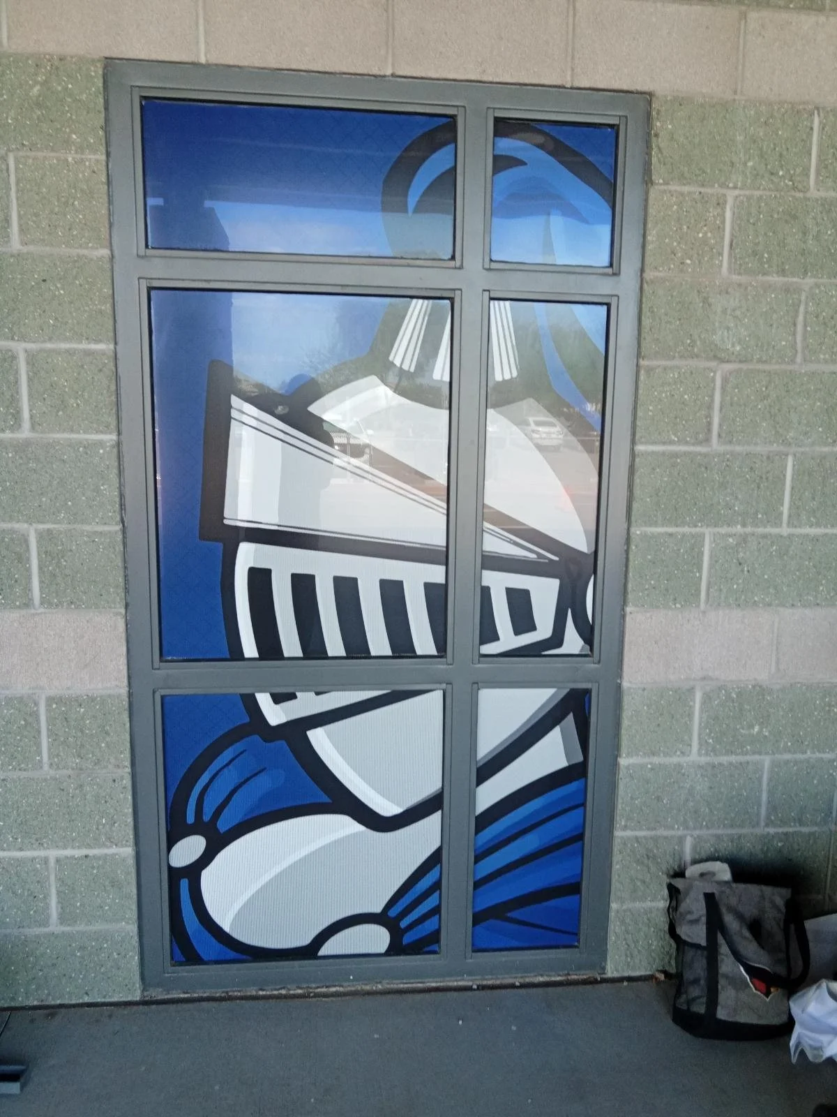 Stencil art of a person wearing a hoodie and a face mask, with a blue background, displayed on a large window divided into six panes, mounted on a brick wall.
