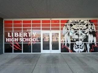 Glass window display with 'Liberty High School' in white letters, a red, white, and blue background, and a large lion mascot on the right side.