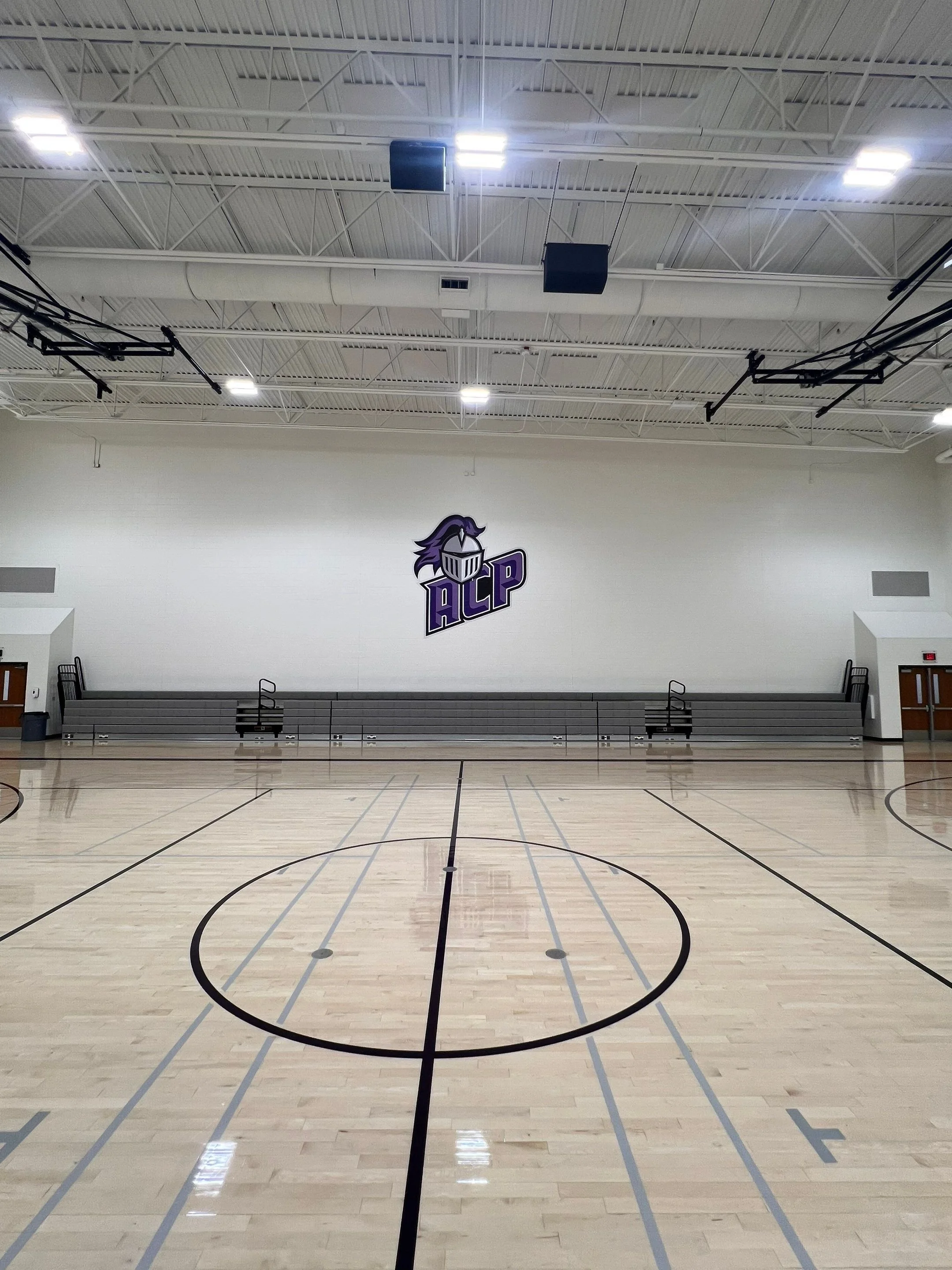 Empty indoor basketball court with a logo of a knight's helmet and the letters 'ACP' on the wall. Bleachers are on both sides of the court.