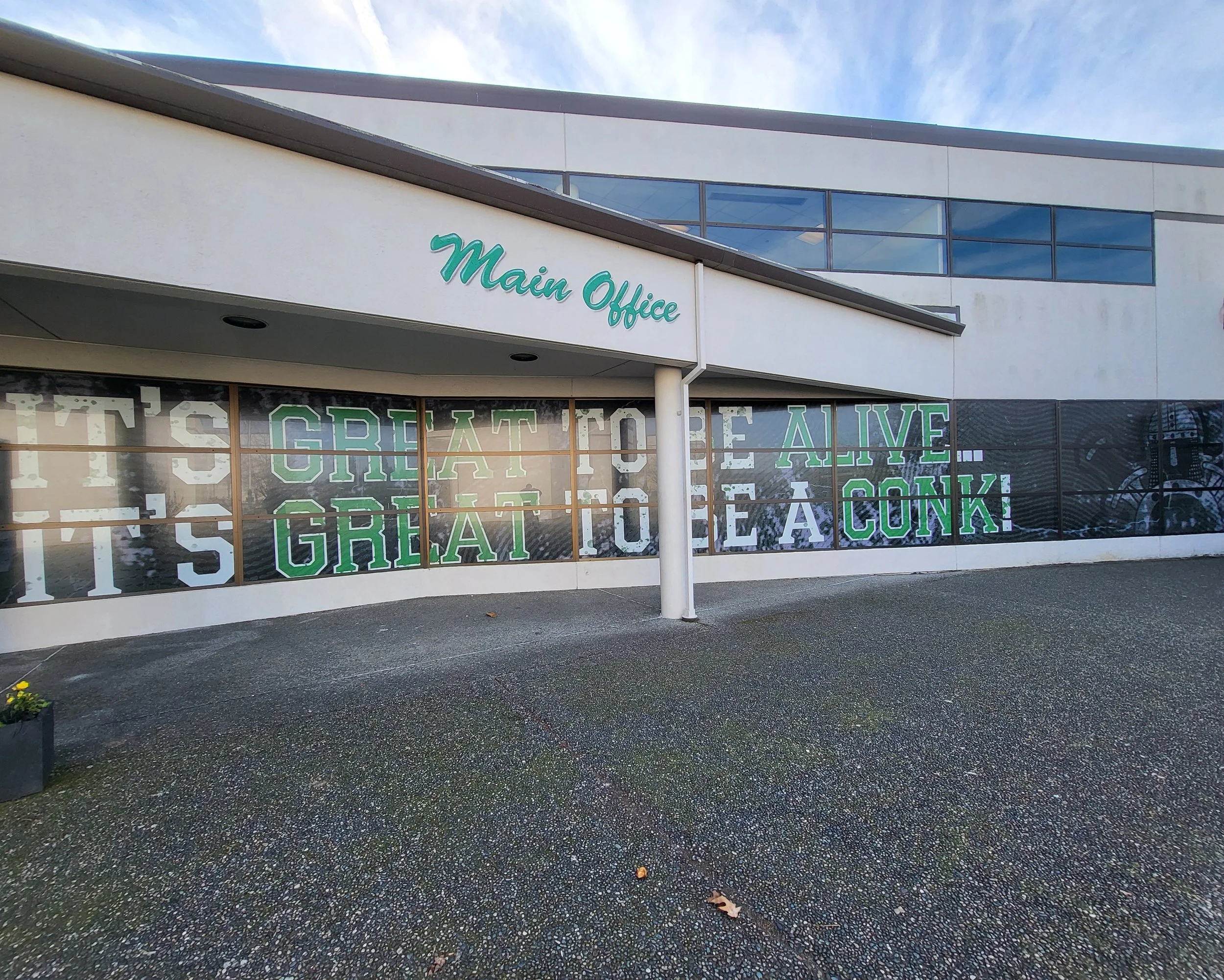 Exterior of a building with a sign reading 'Main Office' on a white wall. The building has large windows and a black awning with large glass storefront windows below that have motivational words like 'GREAT' and 'TOGETHER' visible through them. The g