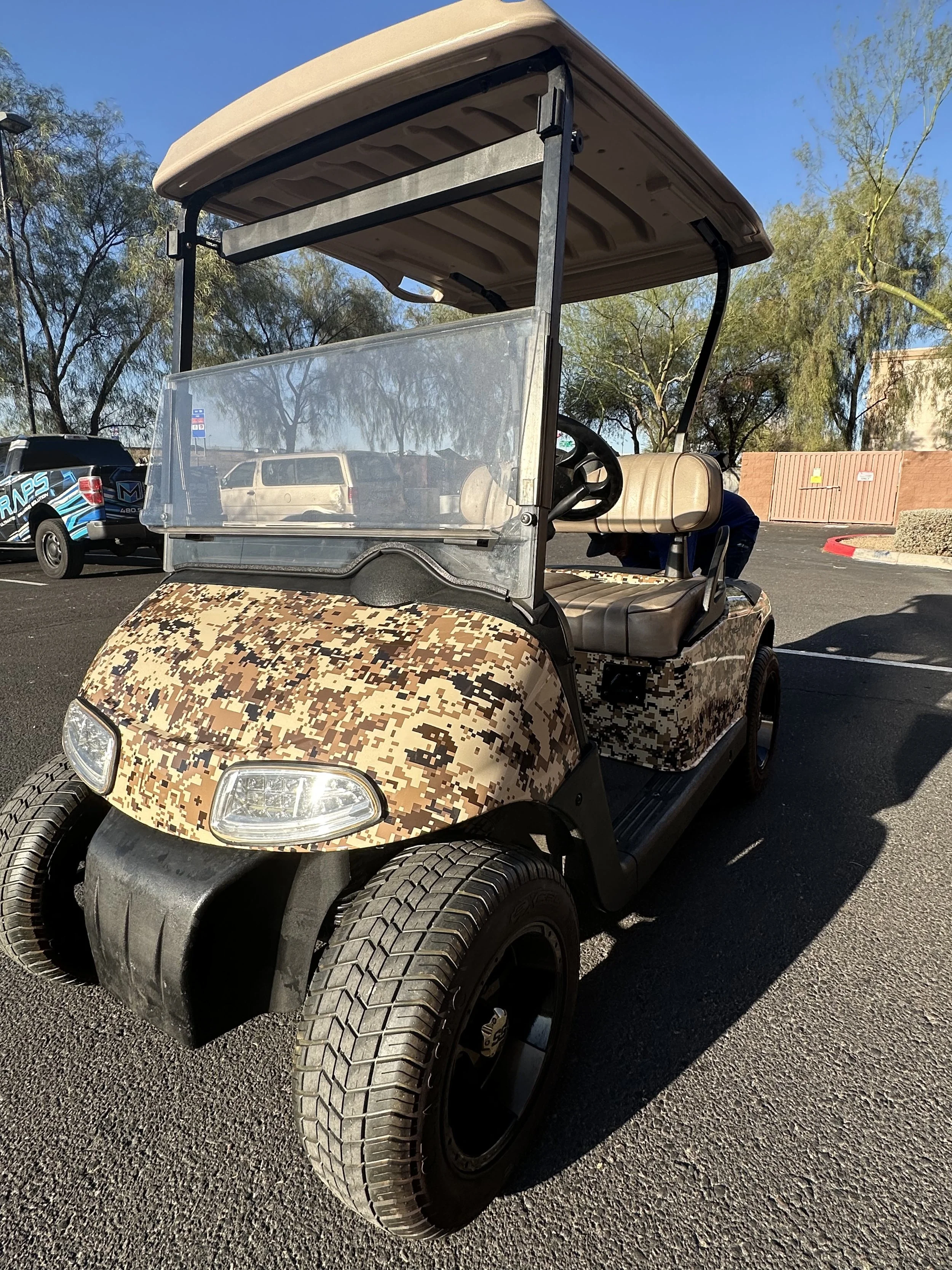A golf cart with desert camouflage pattern on the front, parked in a lot with other vehicles and trees in the background.