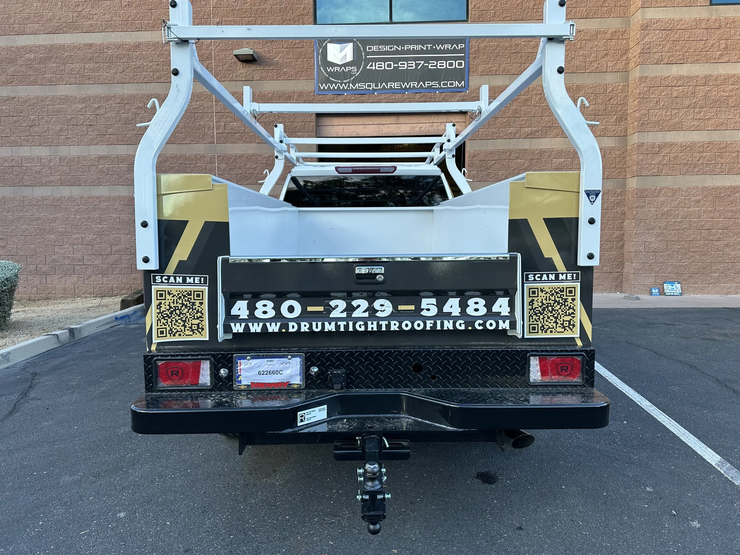 The back of a trailer with signage for MSquare Wraps and contact information, parked on a paved lot in front of a brick building.