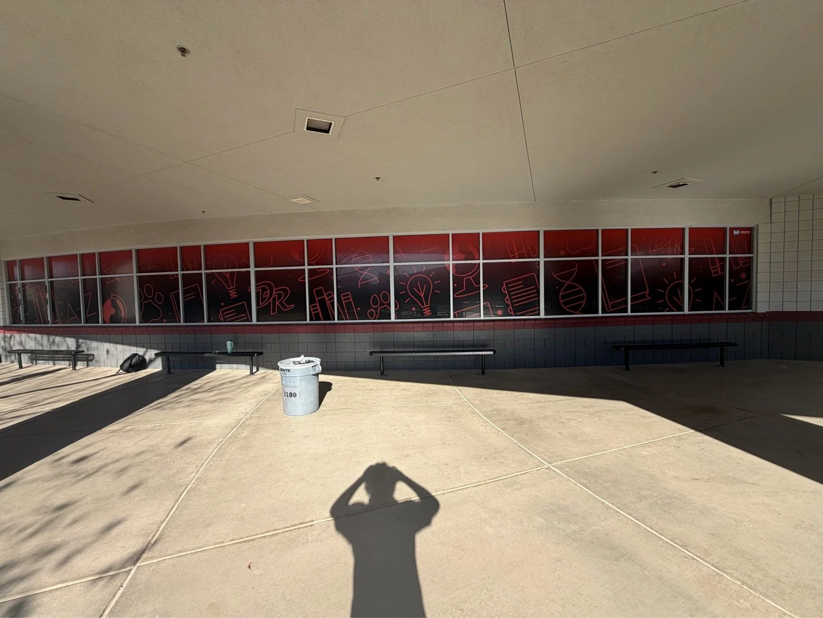An outdoor area with a large tiled wall featuring a red and black graphic design of books, lightbulbs, and scientific symbols. There are three black benches, a trash can, and a backpack on the ground. Shadows are cast on the beige concrete ground, an