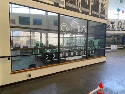 Glass window with a neon sign, logo, and the words 'Hollywood Conquerors,' inside an airport or mall, with a corridor and some artwork on the wall reflecting on the glass.