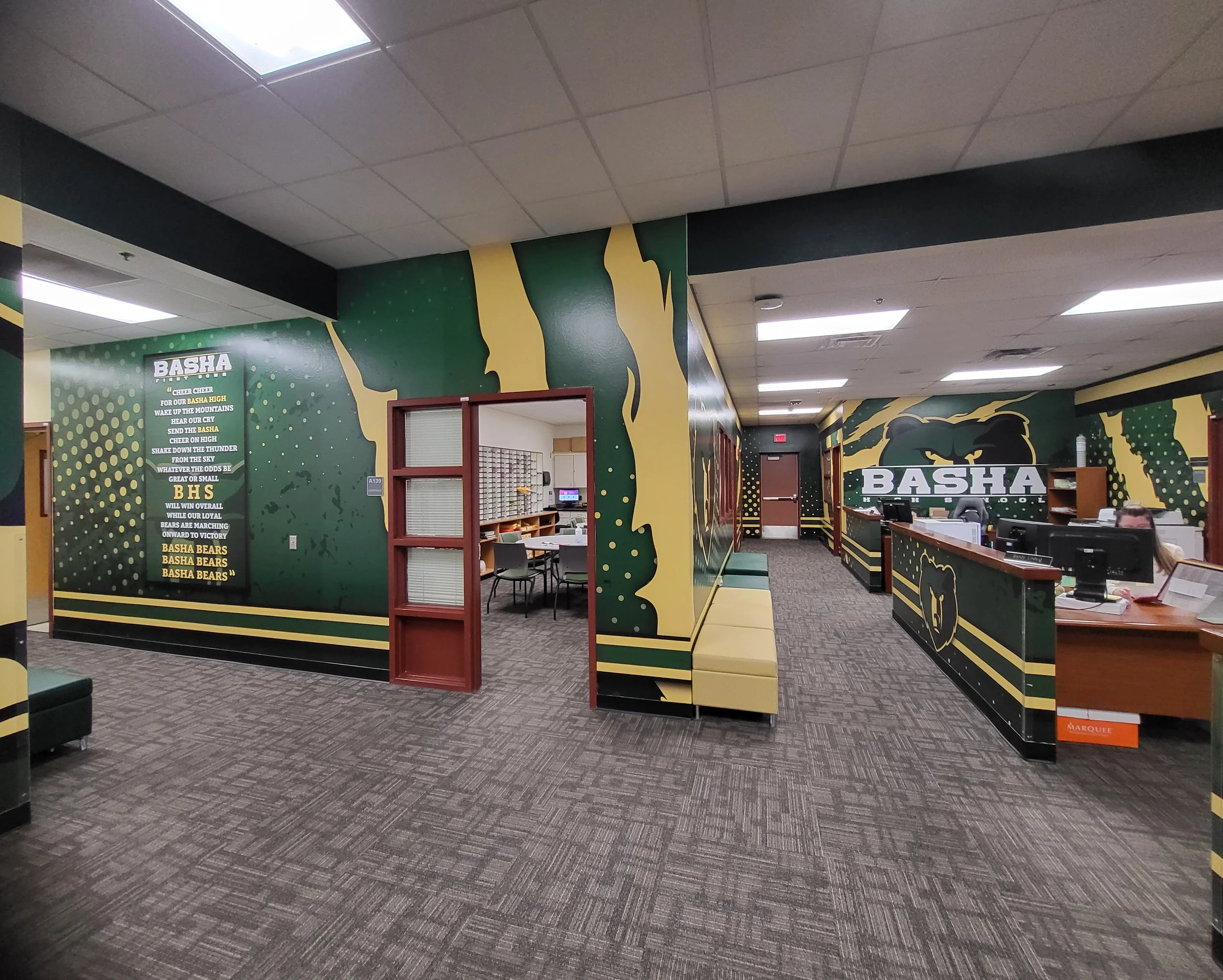 Interior of a school or office with green and yellow wall murals and a reception desk. The ceiling has fluorescent lights and a large skylight.