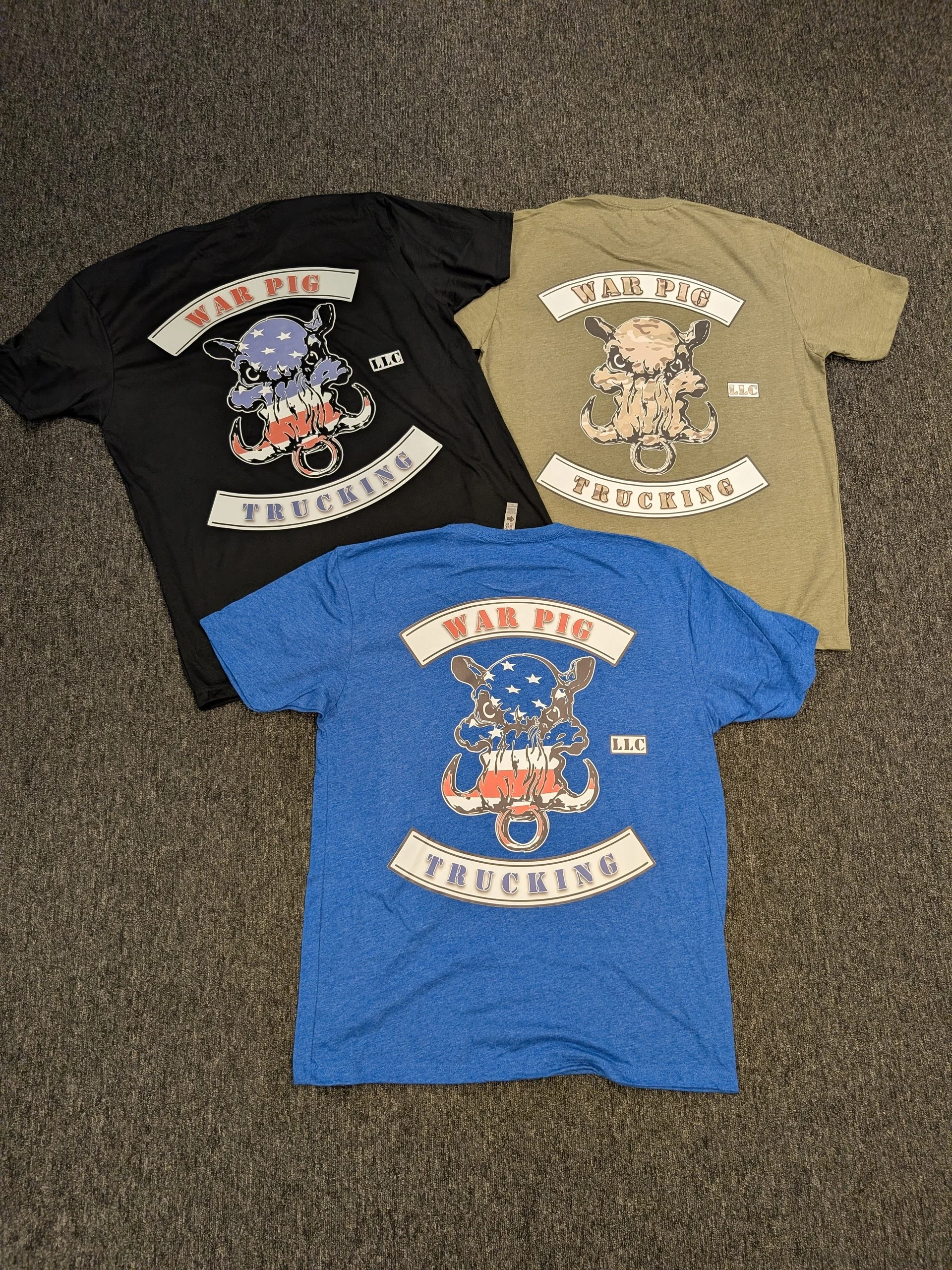Three t-shirts on a carpeted floor, each with a graphic design featuring a stylized skull with horns and the words "WAR PIG TRUCKING" and "LLC".