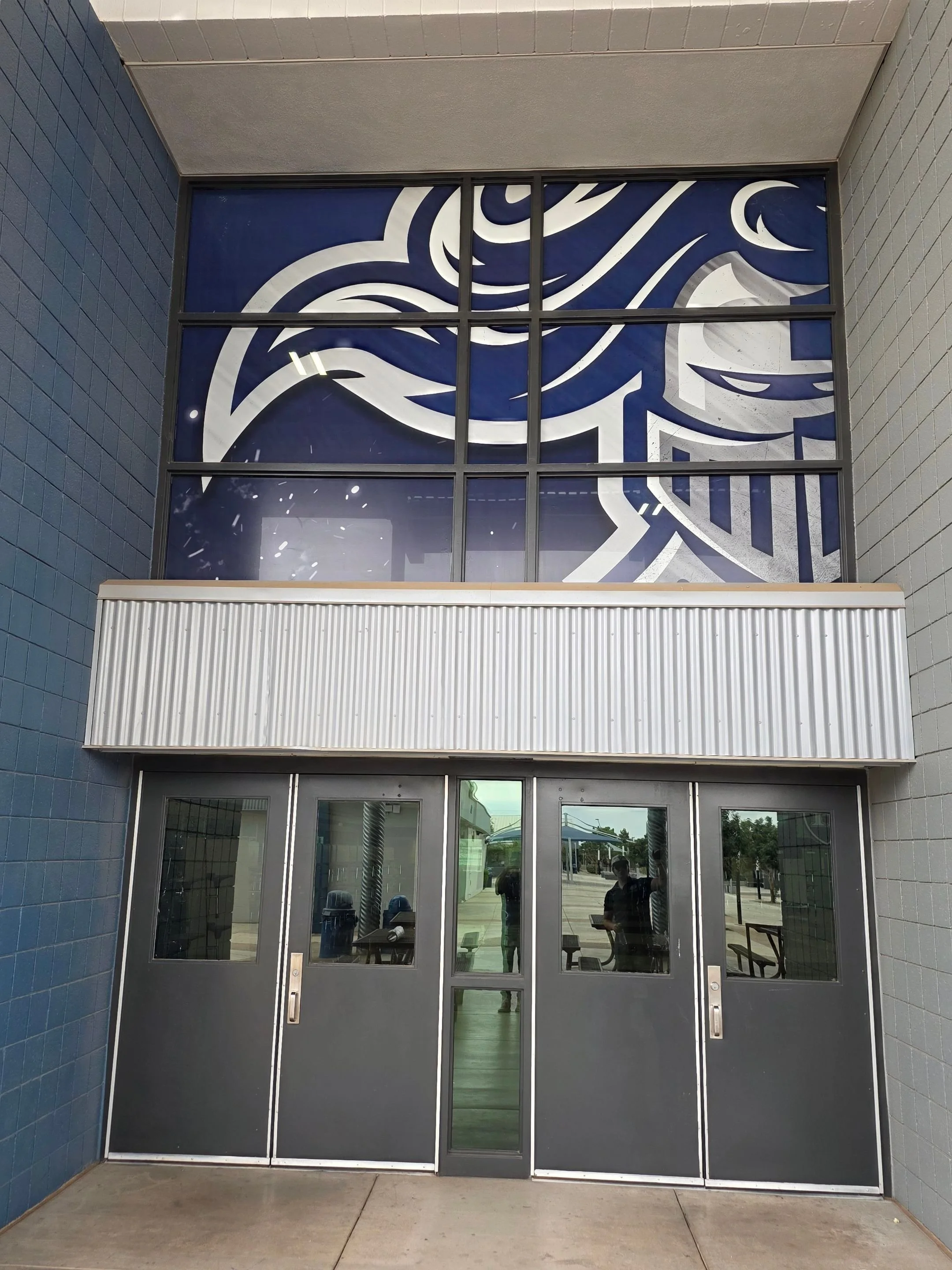 Exterior view of a building entrance with glass double doors and a large decorative window above. The window features abstract, white-lined patterns on a blue background, seen through the transparent glass. The surrounding walls are tiled in blue and