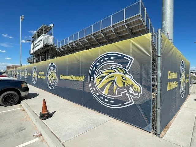 Construction fence with logos of the school and the slogan 'Commitment' in yellow, next to a parking lot and a sports press box.