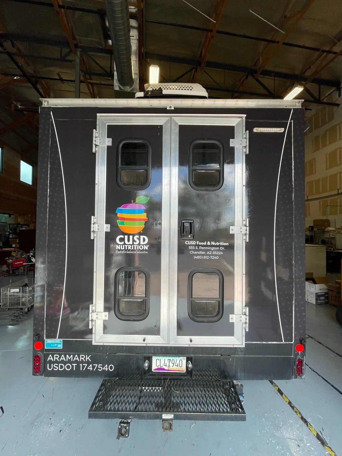 The back of a black food truck with a colorful apple logo and the words 'CUSD Nutrition' and contact information, parked inside a warehouse or garage.