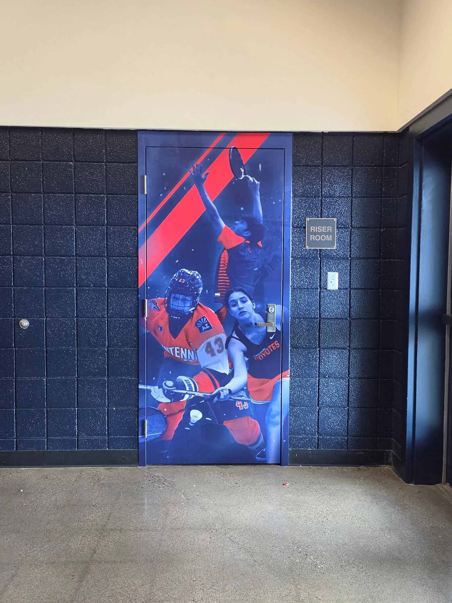 A door with a poster of two hockey players, one in a red uniform with the number 43 and a goalie mask, and the other in a black uniform with a baton, with a black tiled wall around it and a small sign that says 'Riser Room'.