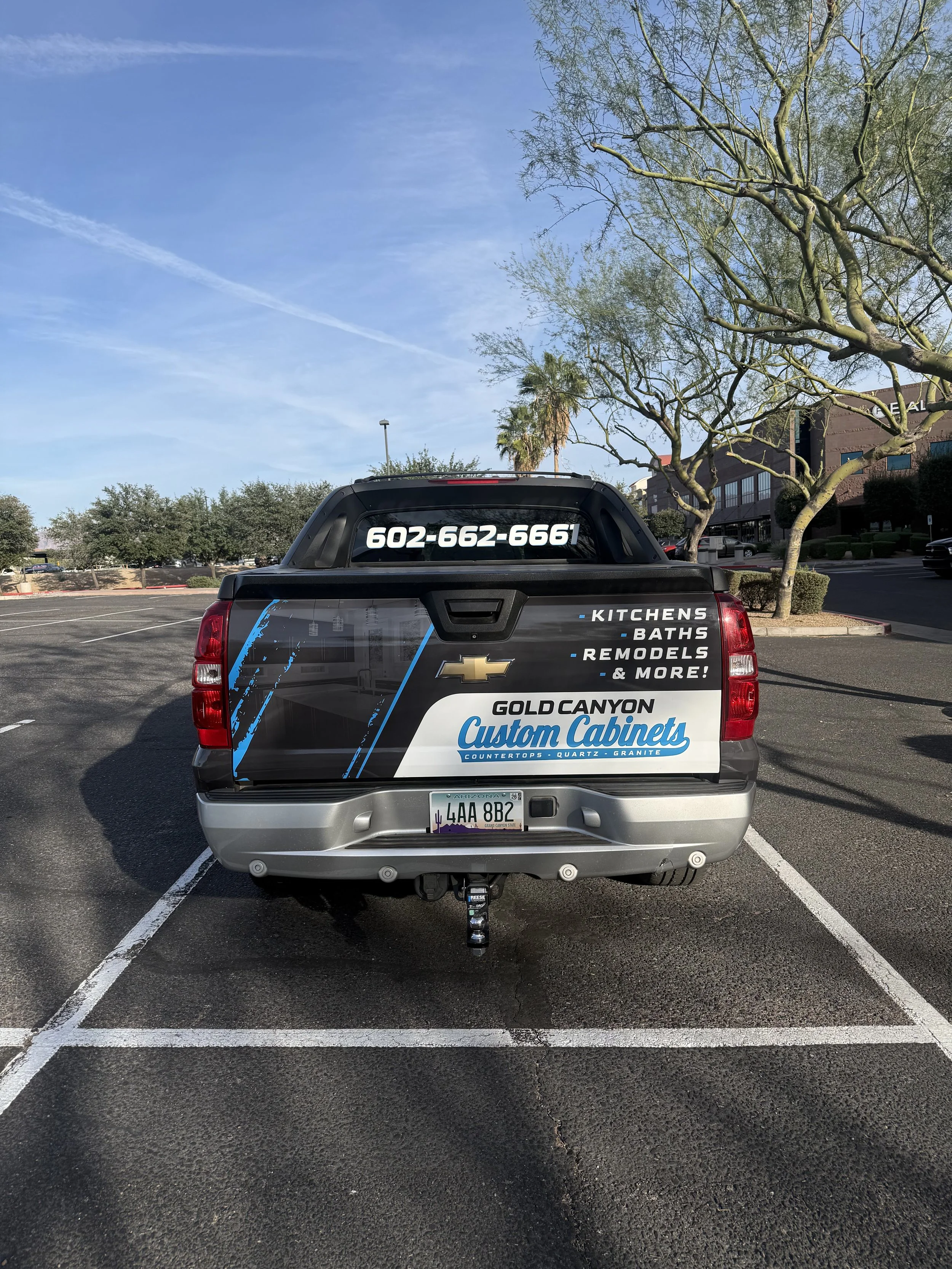 A black pickup truck parked in a parking lot with a large advertisement on the back, displaying a phone number and services including kitchens, baths, remodels, and countertops. The truck has a visible Arizona license plate and is parked among other 