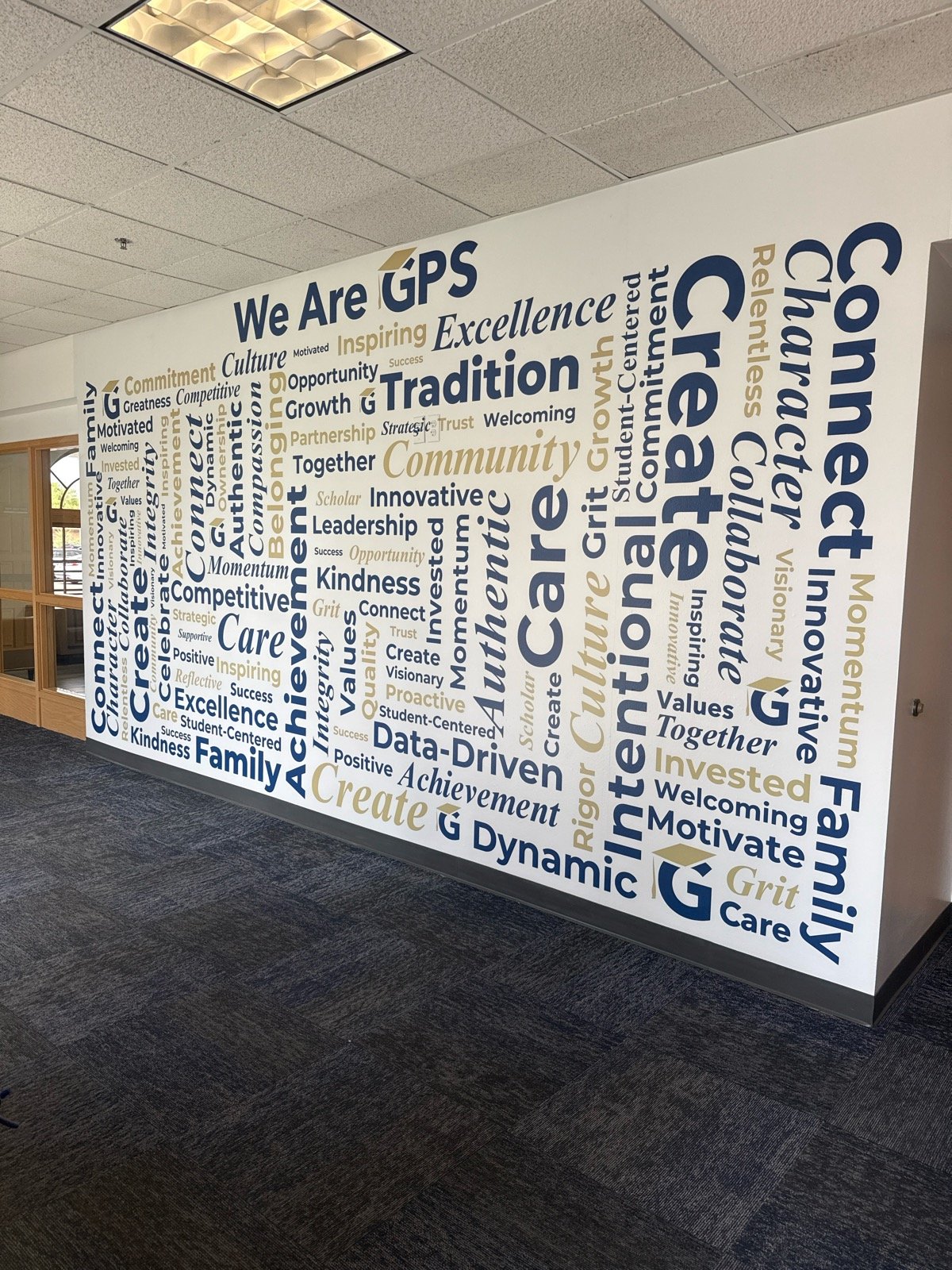 Interior of an office space featuring a large wall mural with words like 'We Are GPS', 'Create', 'Community', and 'Innovation' in varying sizes and colors, emphasizing the organization's values and mission.