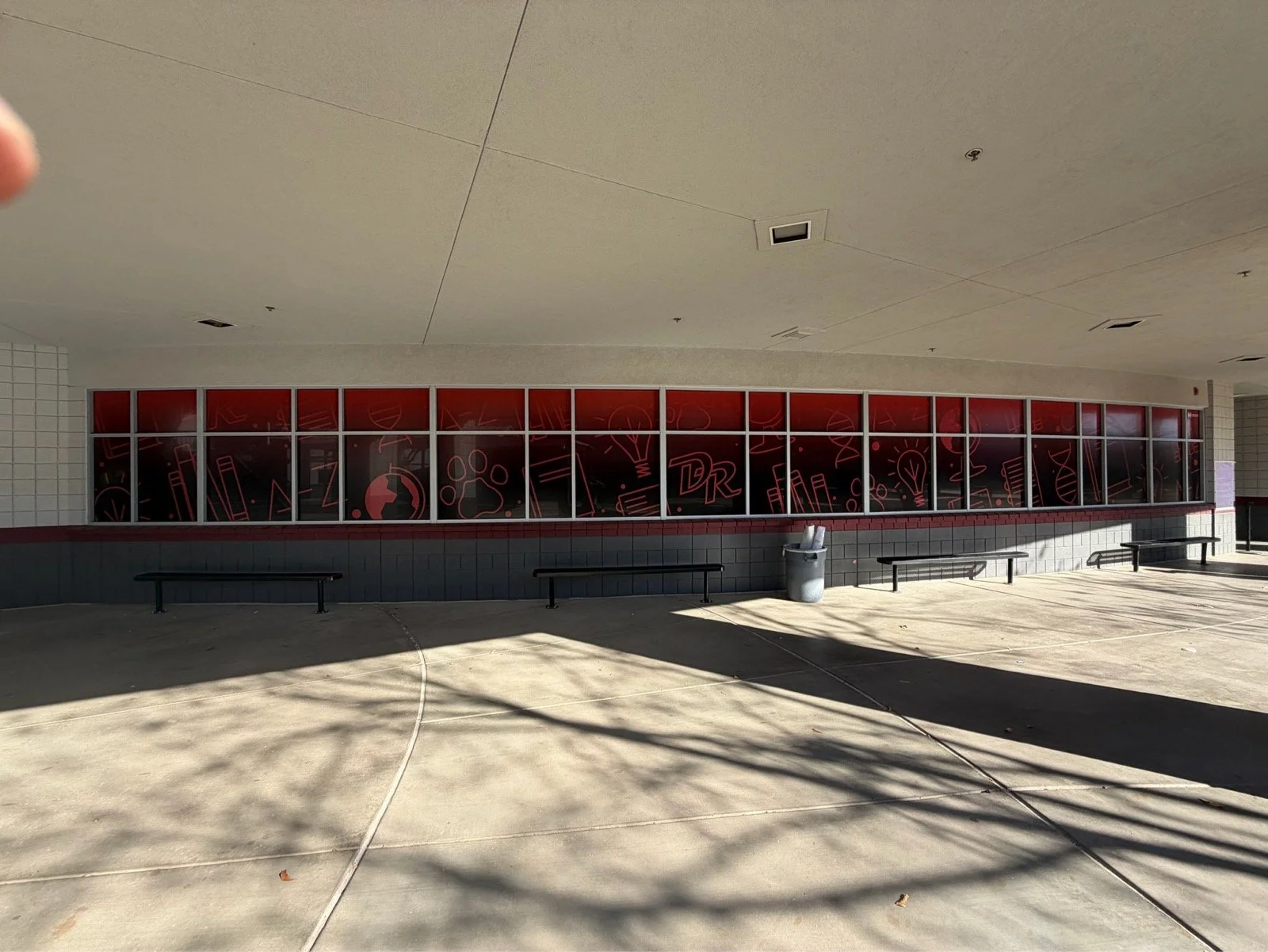 A curved building with windows featuring red backgrounds and science-themed white line art, including test tubes, DNA helixes, and light bulbs, and benches underneath.