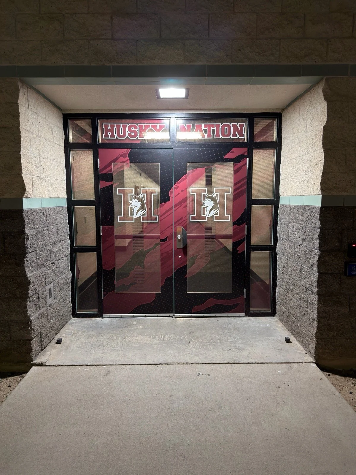 Double doors with Huskies logo and large letter 'H' on each door, framed by brick and concrete walls, with an overhead light illuminating the entrance.