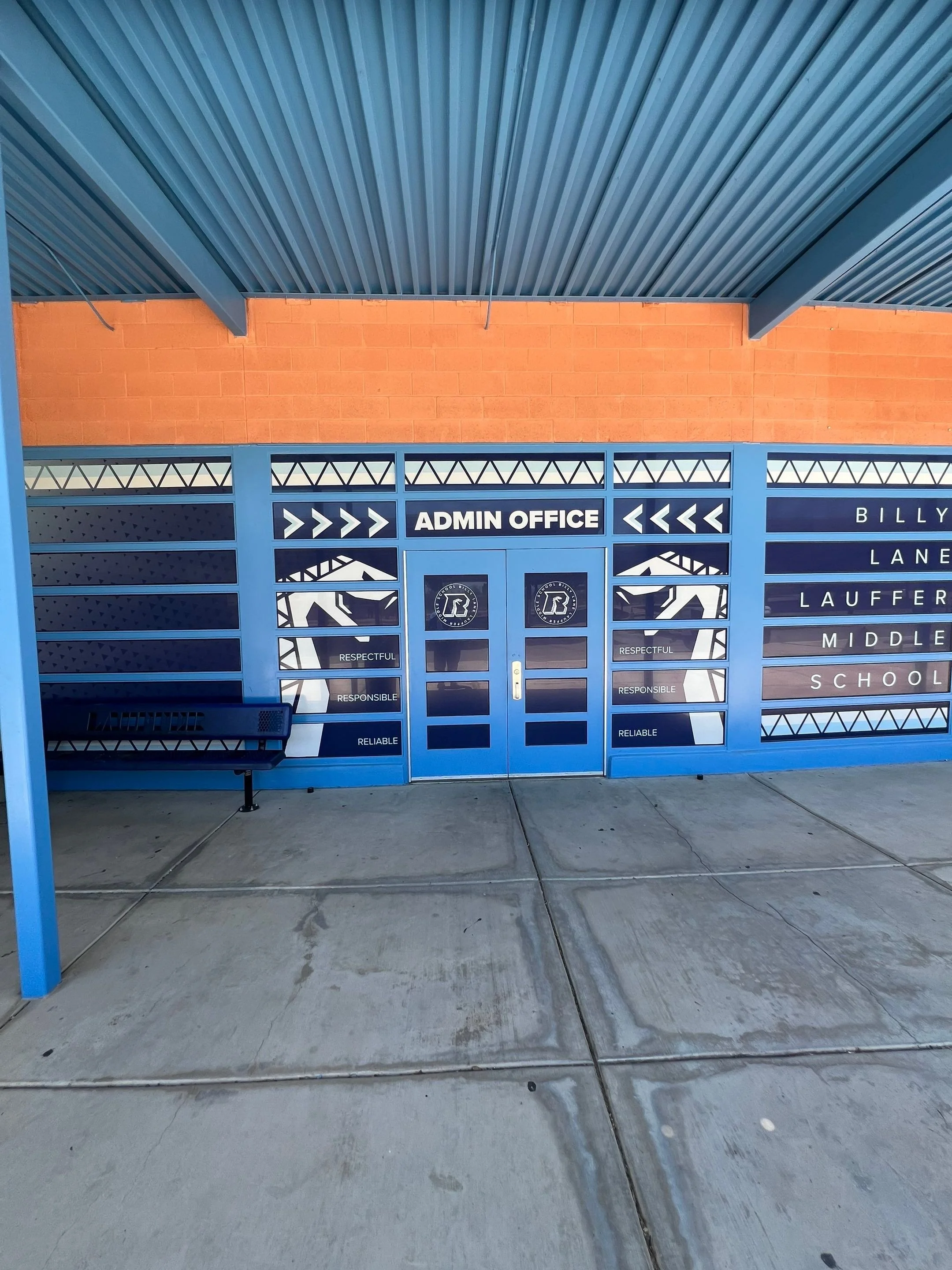 School entrance with blue gate, signage indicating 'Admin Office' and various school levels, a black bench, and blue metal roof over a concrete sidewalk.
